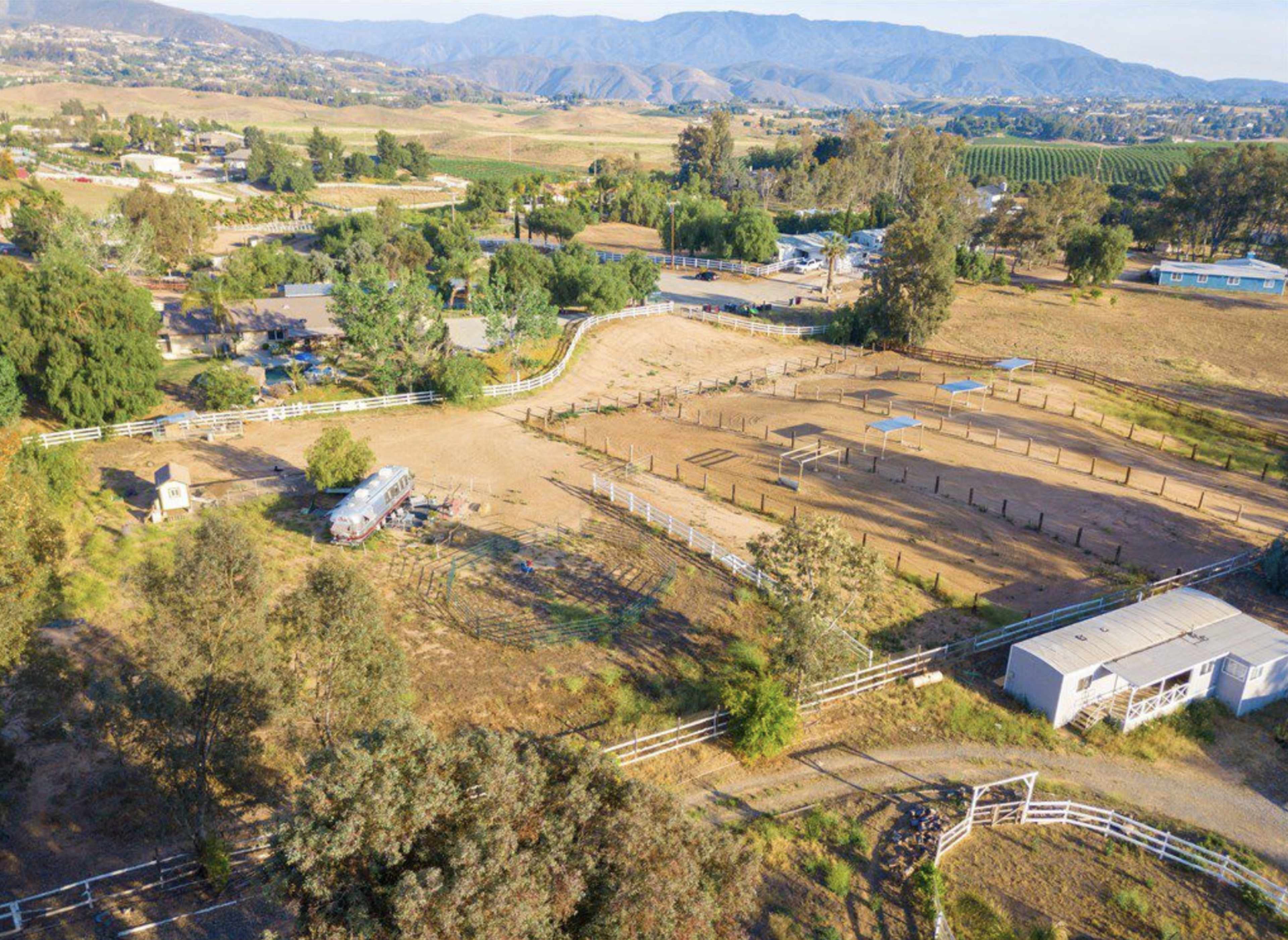 The image shows a rural landscape featuring an equestrian area with several paddocks, surrounded by trees and hills in the background.