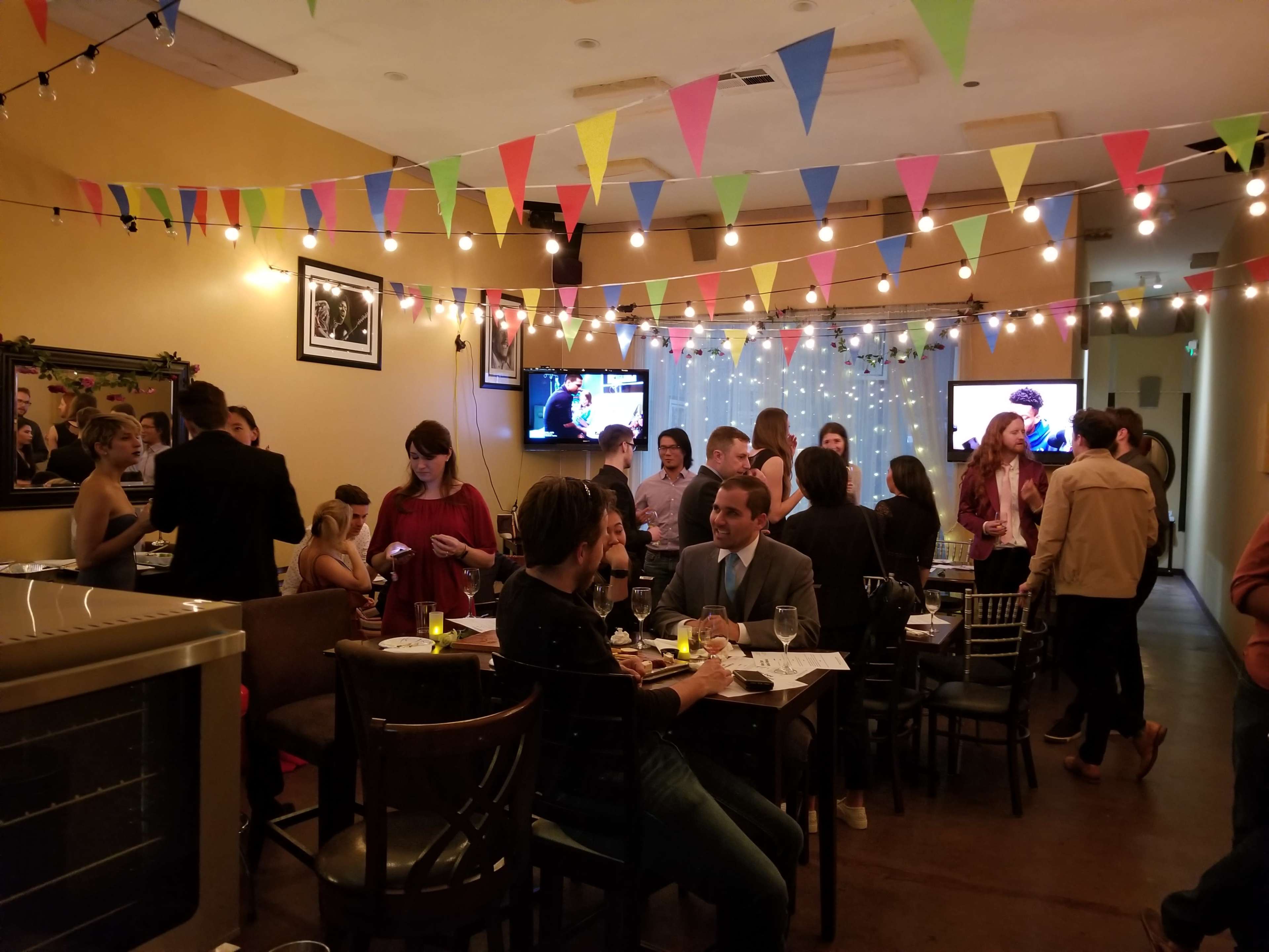 A lively social gathering takes place in a warmly lit room decorated with colorful bunting and string lights, featuring groups of people engaged in conversation.