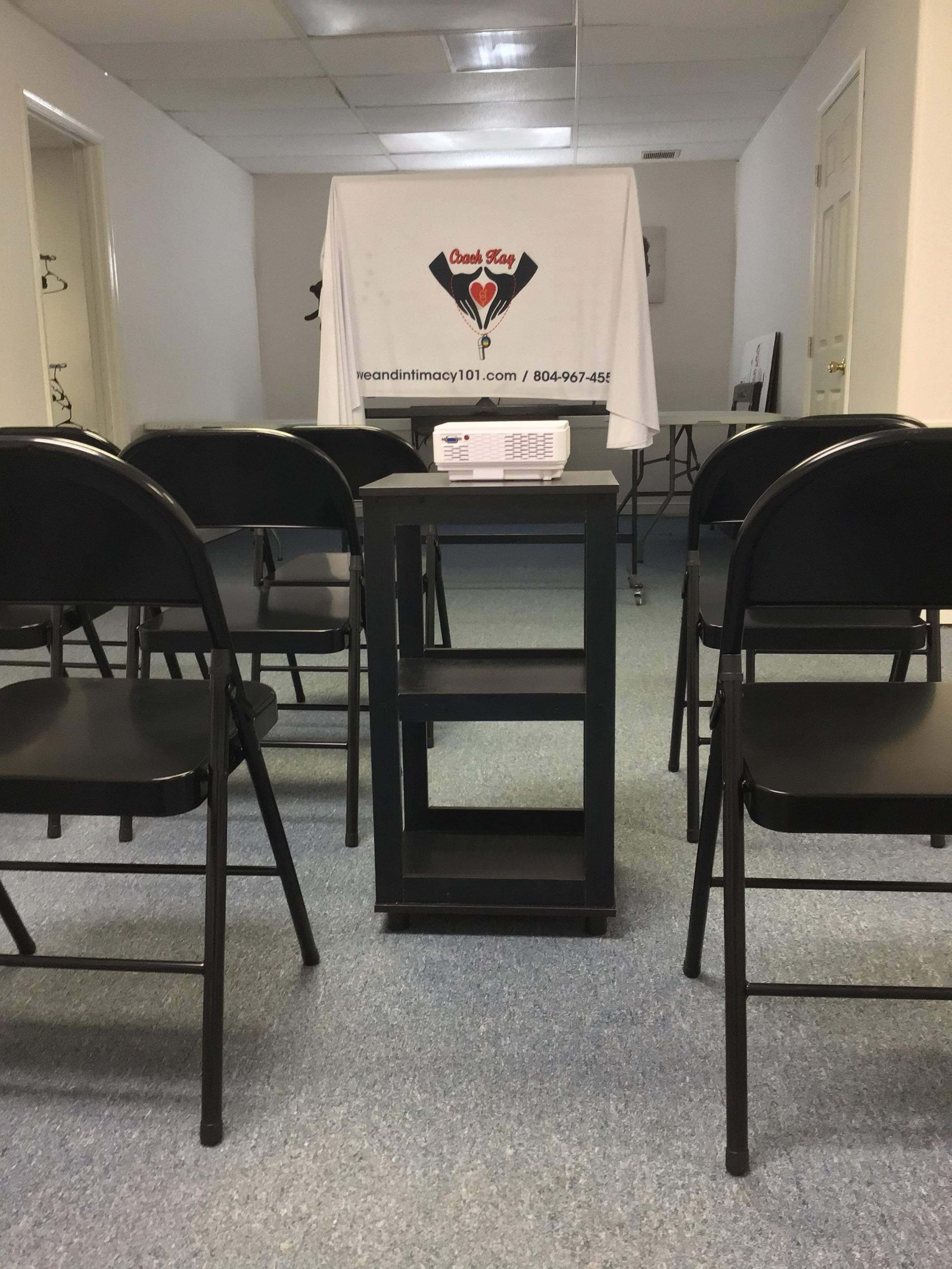 A small meeting room is set up with plastic chairs arranged around a table that holds a projector, facing a screen covered by a white sheet.