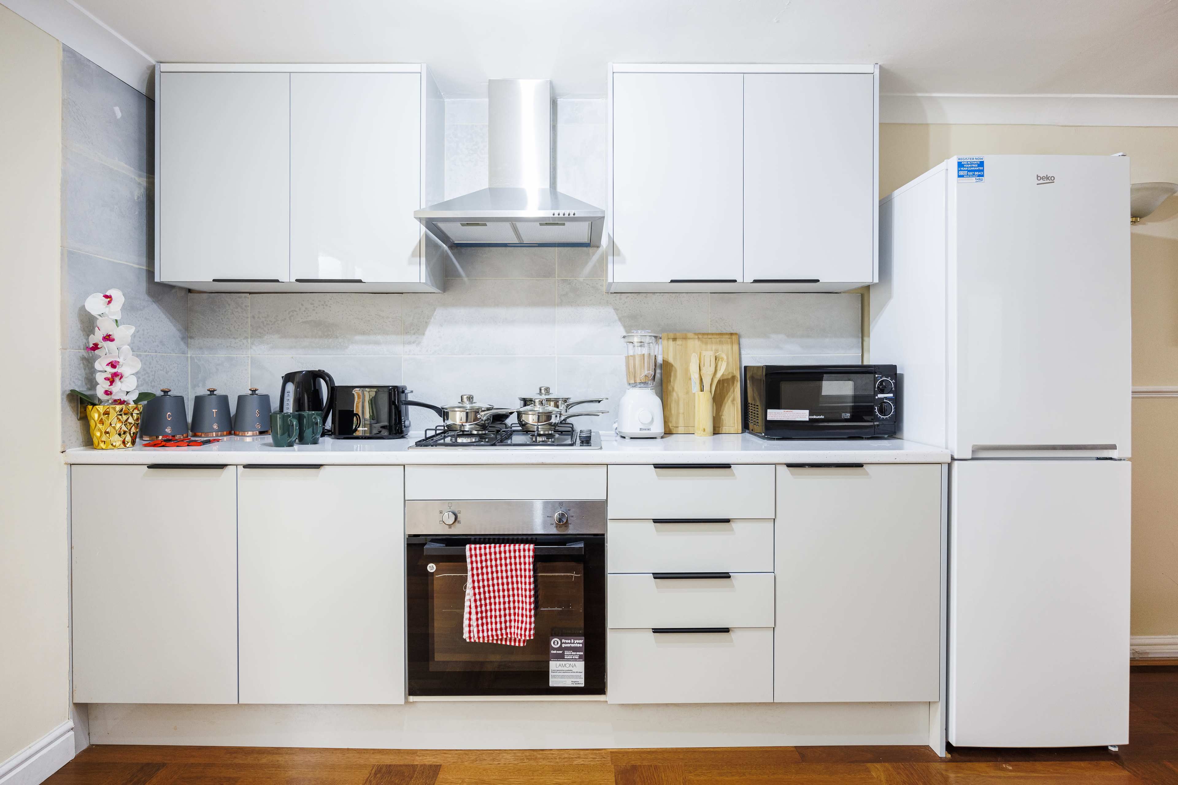 A modern kitchen featuring white cabinetry, a stovetop with an exhaust hood, a microwave, a refrigerator, and various kitchen appliances neatly arranged on the countertop.