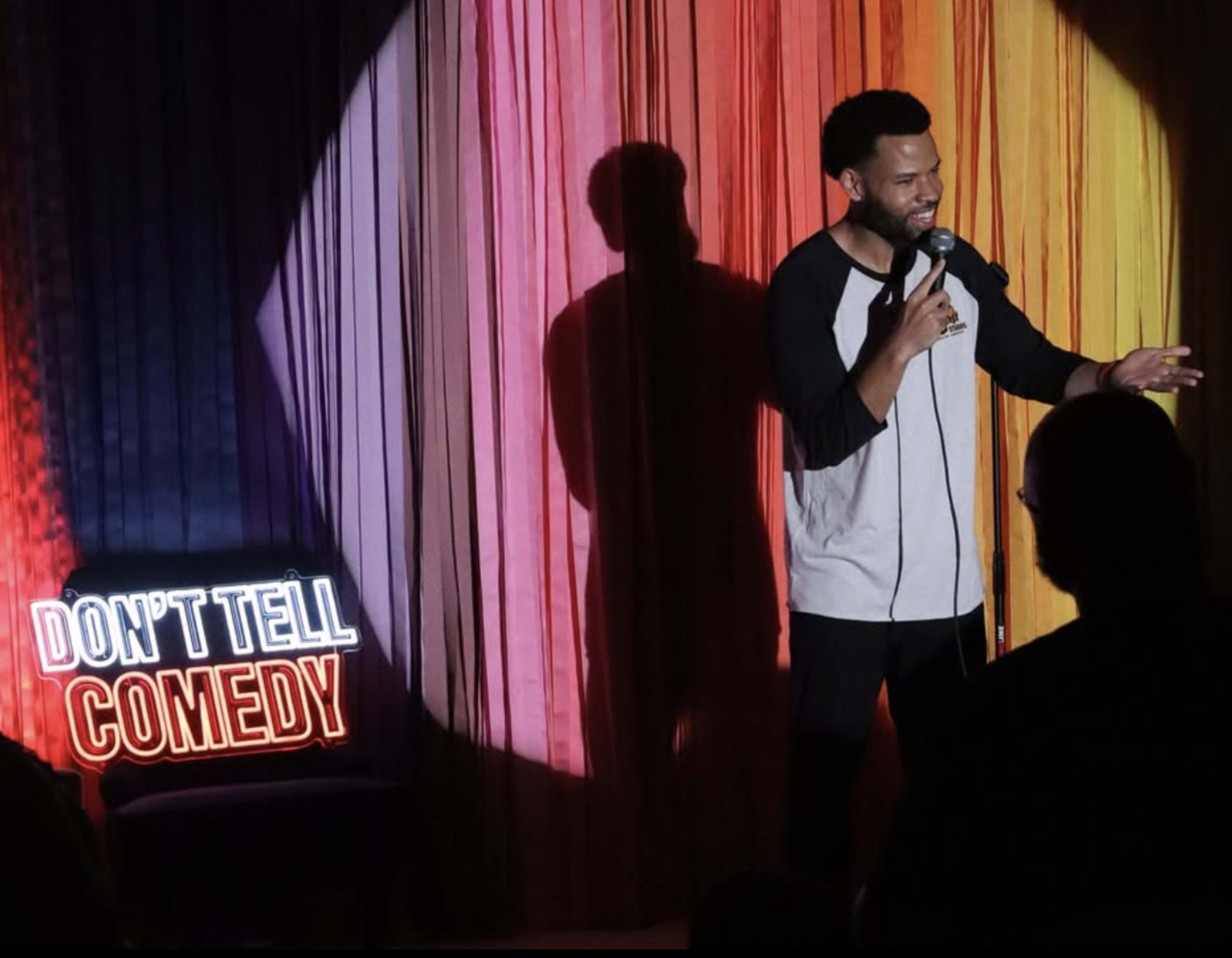 A comedian stands on stage with a microphone, performing in front of an audience while illuminated by colorful curtains and a neon sign that reads "DON'T TELL COMEDY."