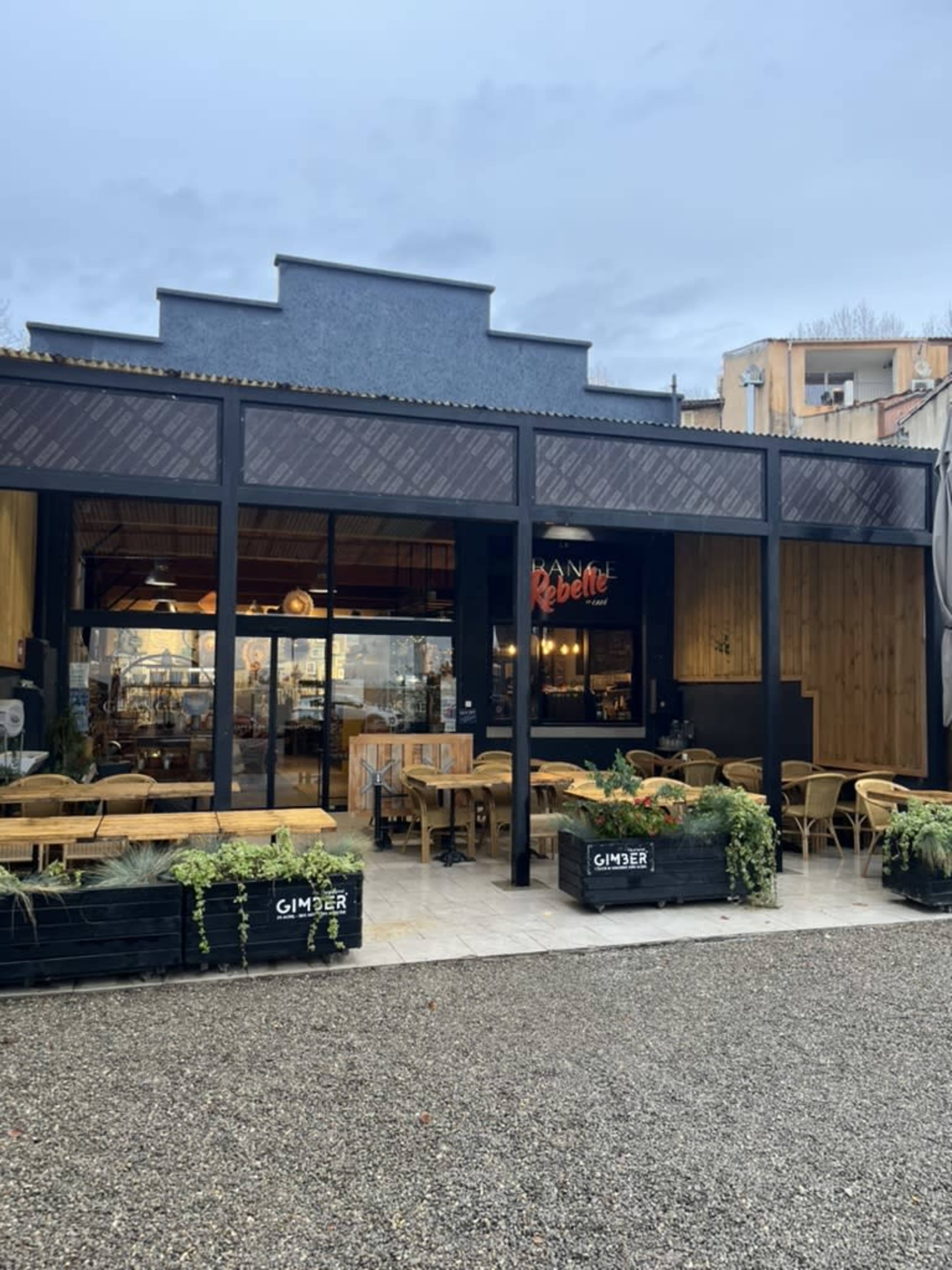The image shows a restaurant with a wooden exterior, large glass windows, and outdoor seating surrounded by potted plants.