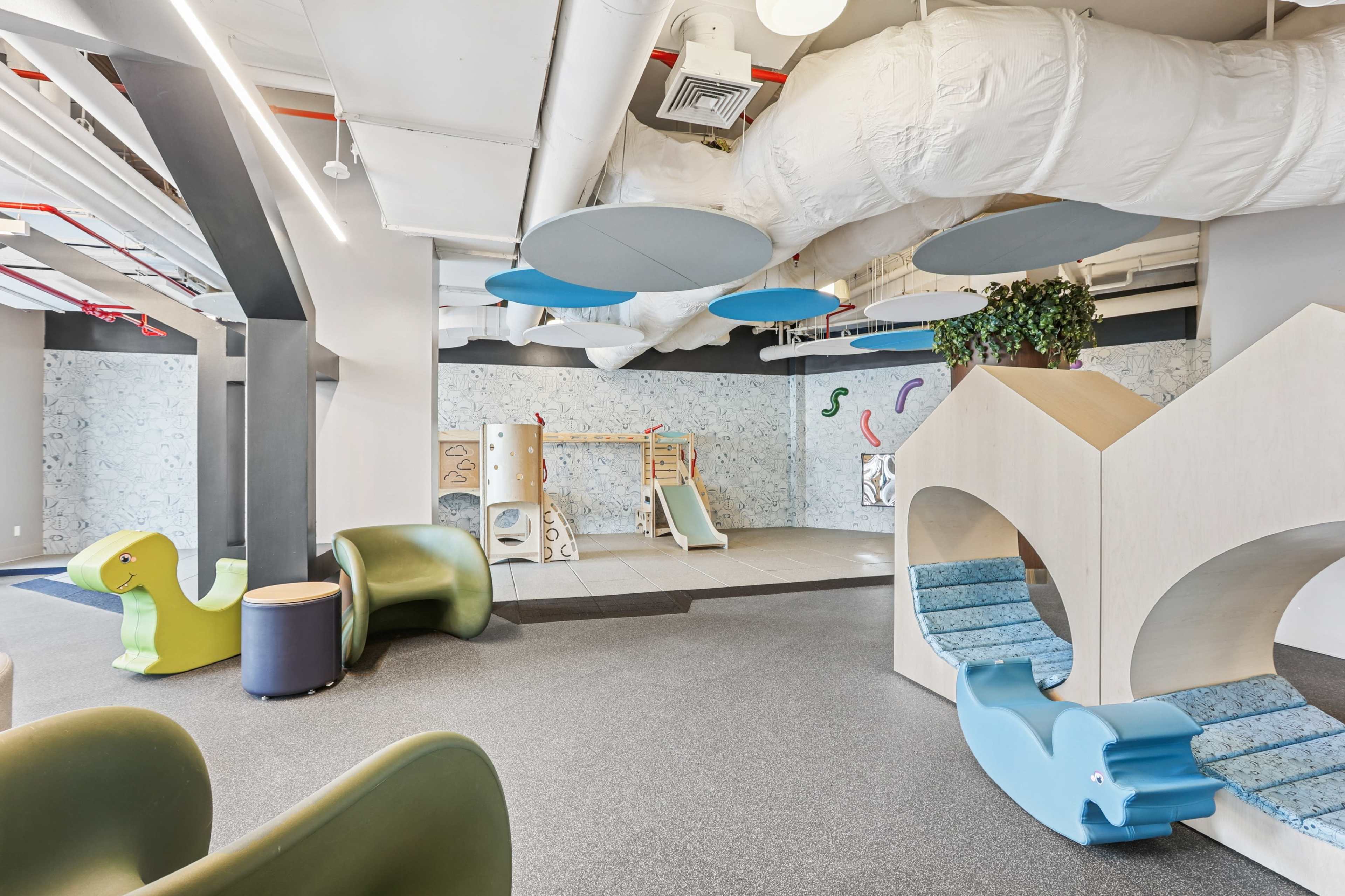 The image shows a brightly lit, modern play area with various shapes of furniture, a climbing structure, and soft flooring.