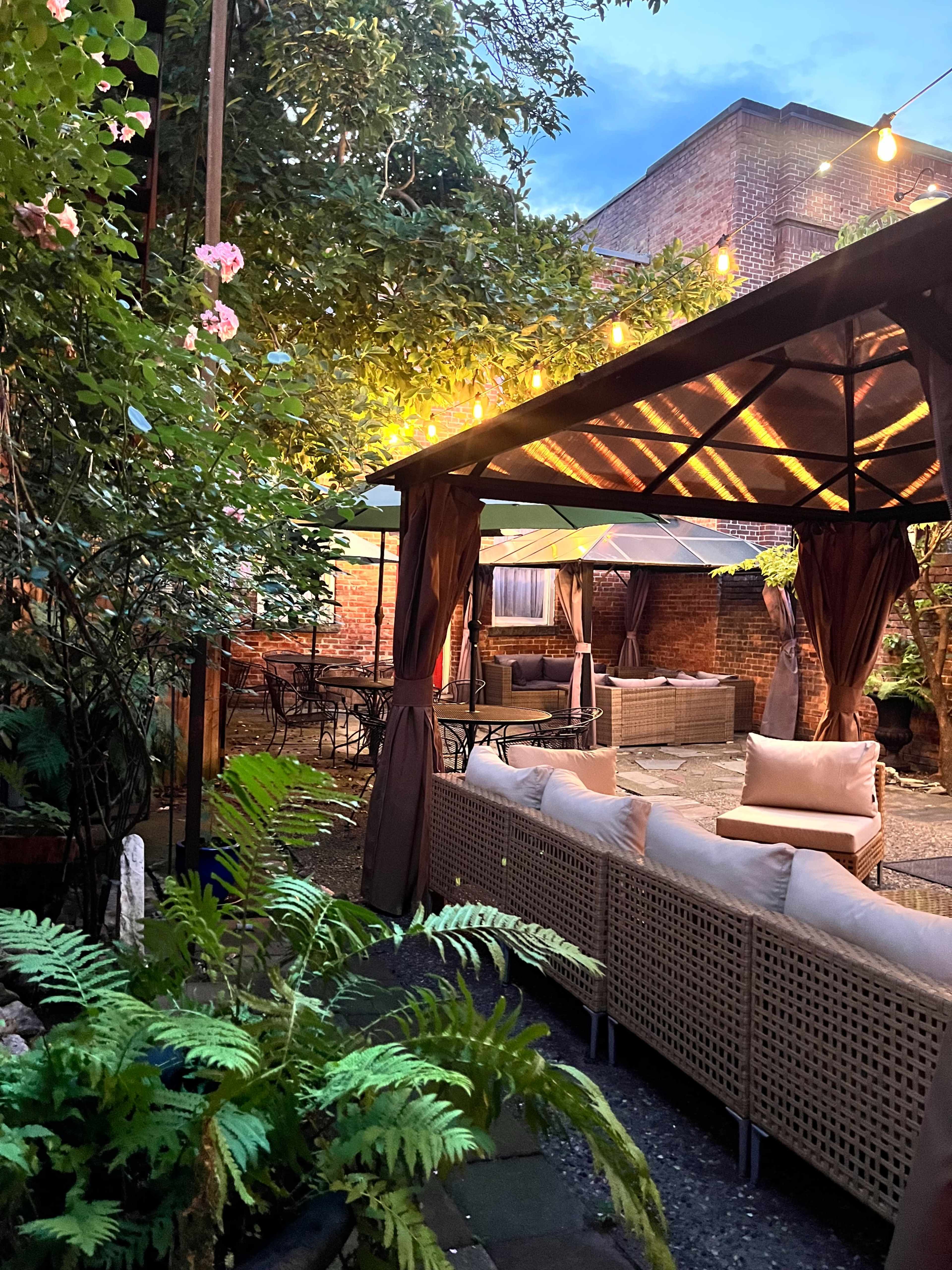 The image shows a cozy outdoor seating area adorned with string lights, surrounded by lush greenery and flowers.