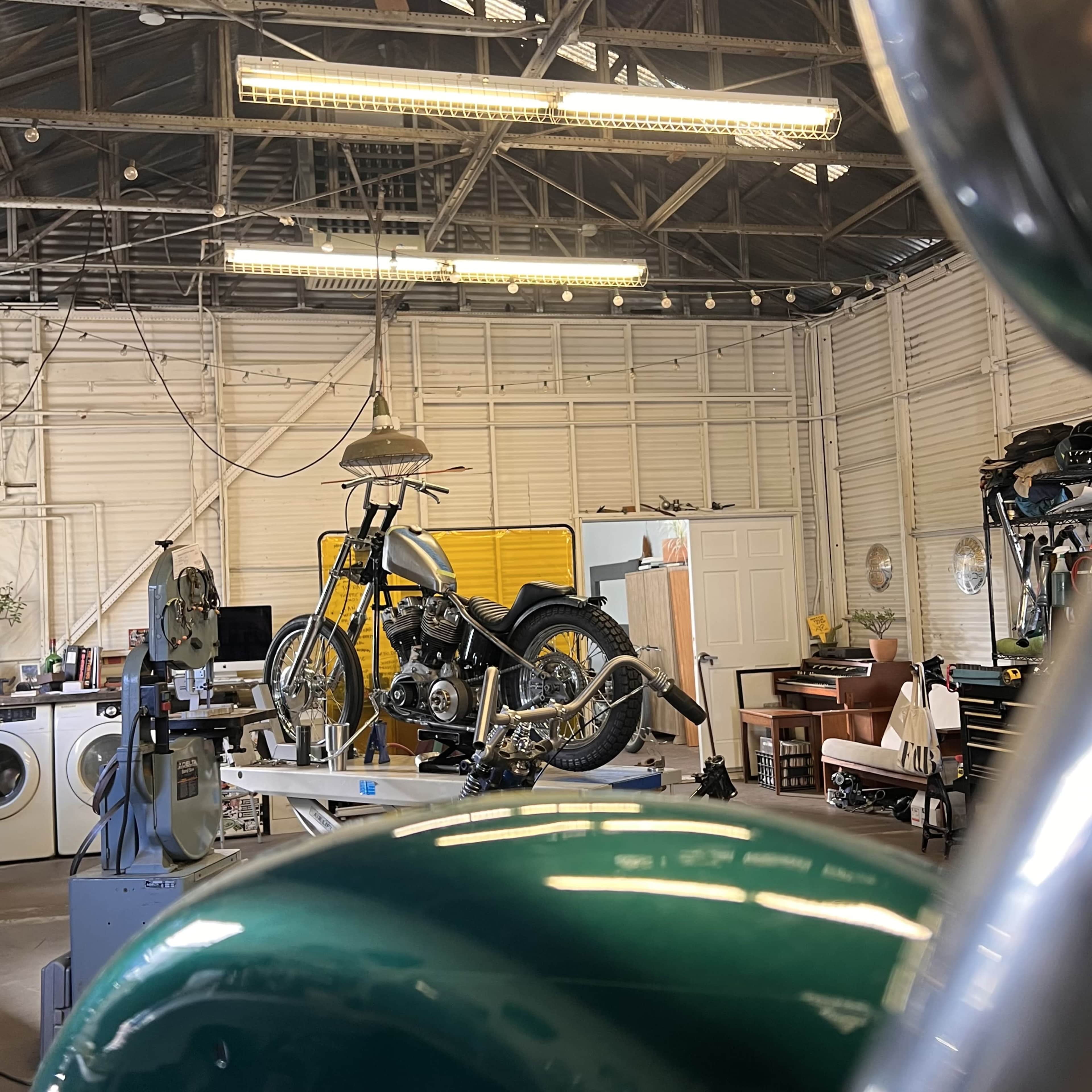 The image shows a motorcycle partially assembled on a workbench in a garage filled with tools and equipment.