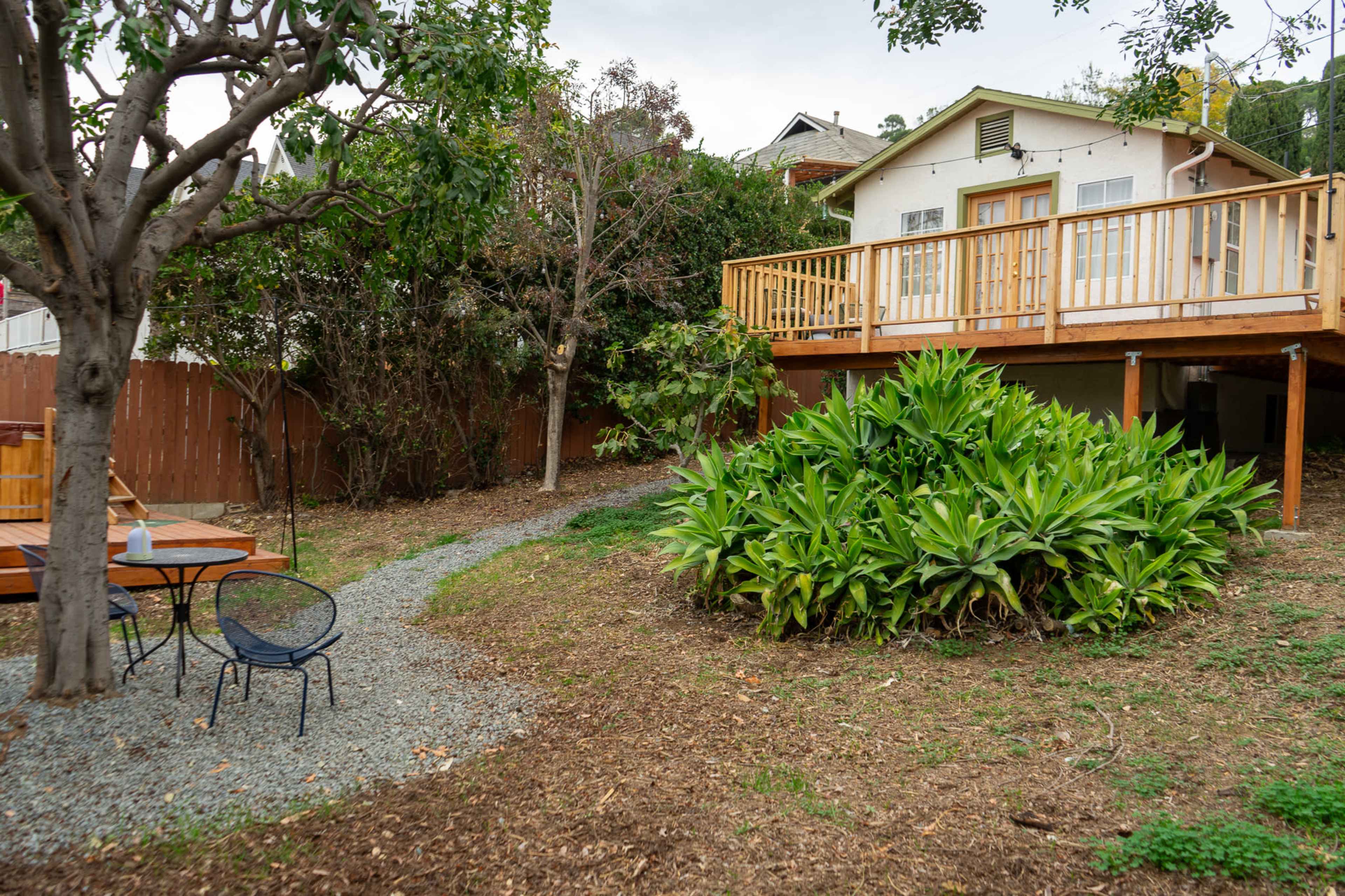 A wooden house on stilts is surrounded by greenery and a gravel path, leading to a small seating area with a table and chair.