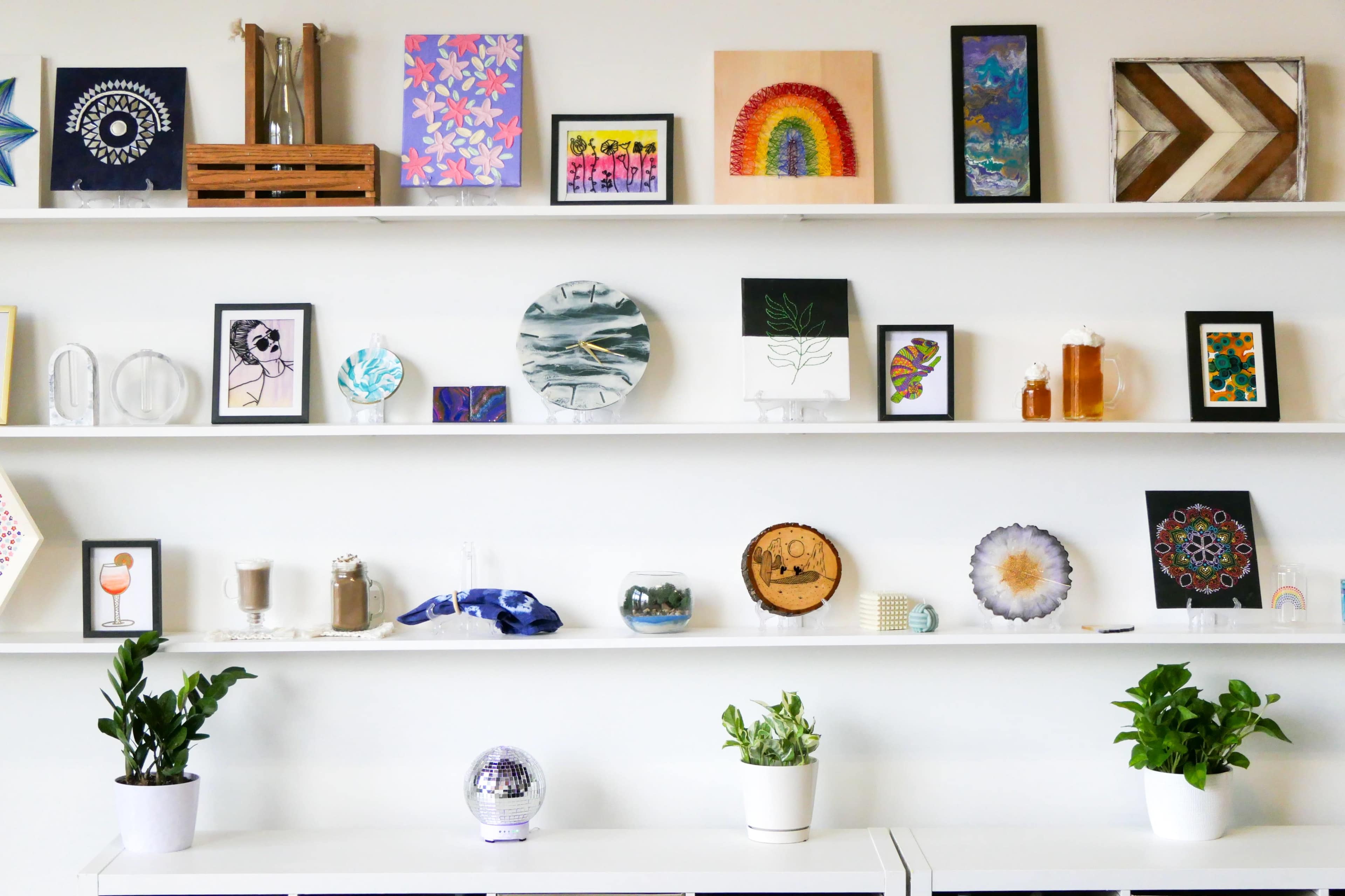 The image shows a series of decorative artworks and plants displayed on multiple shelves against a white wall.