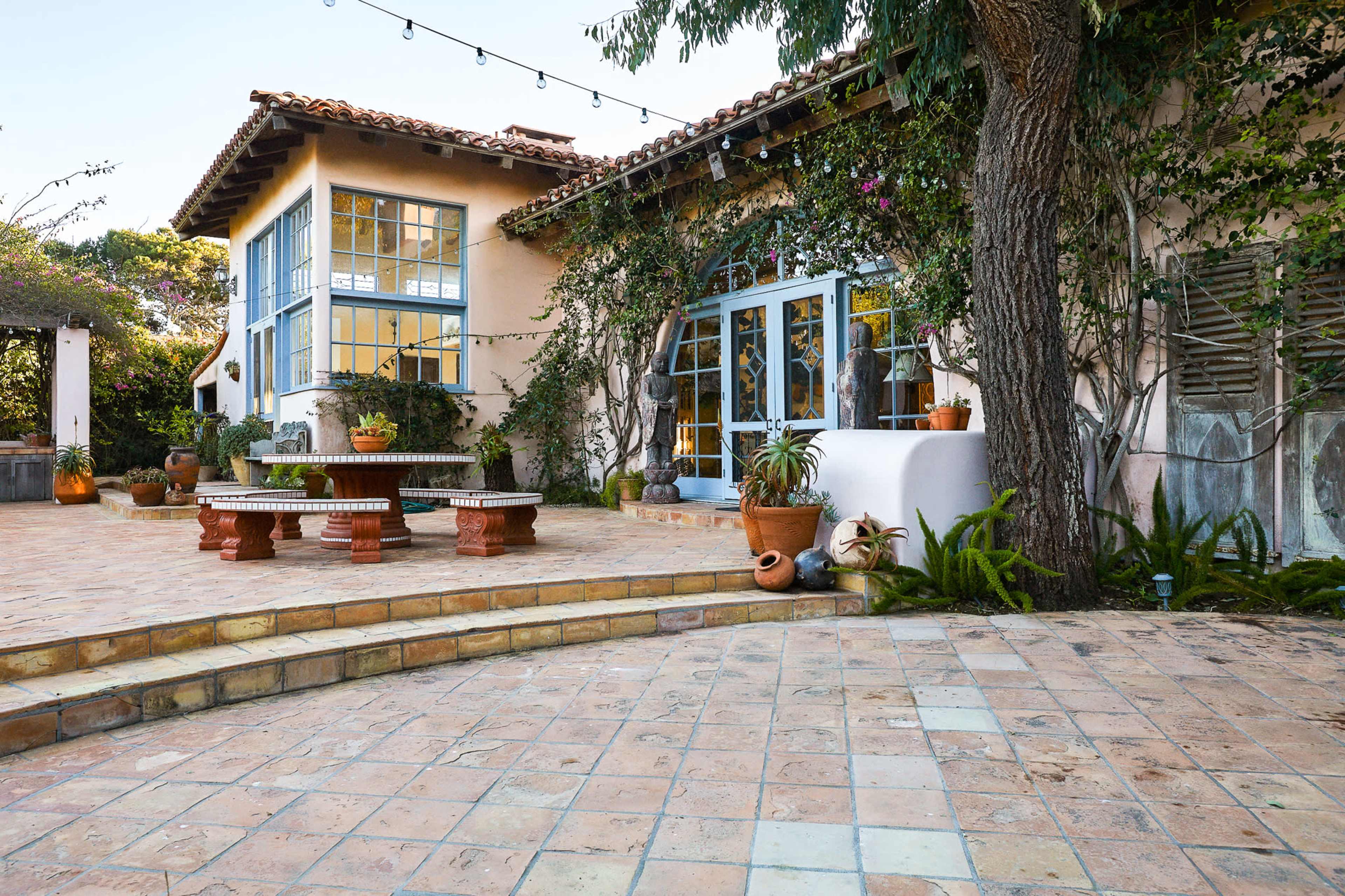A courtyard with a tiled patio featuring potted plants and a rustic wooden bench adjacent to a house with blue-framed windows and flowering vines.