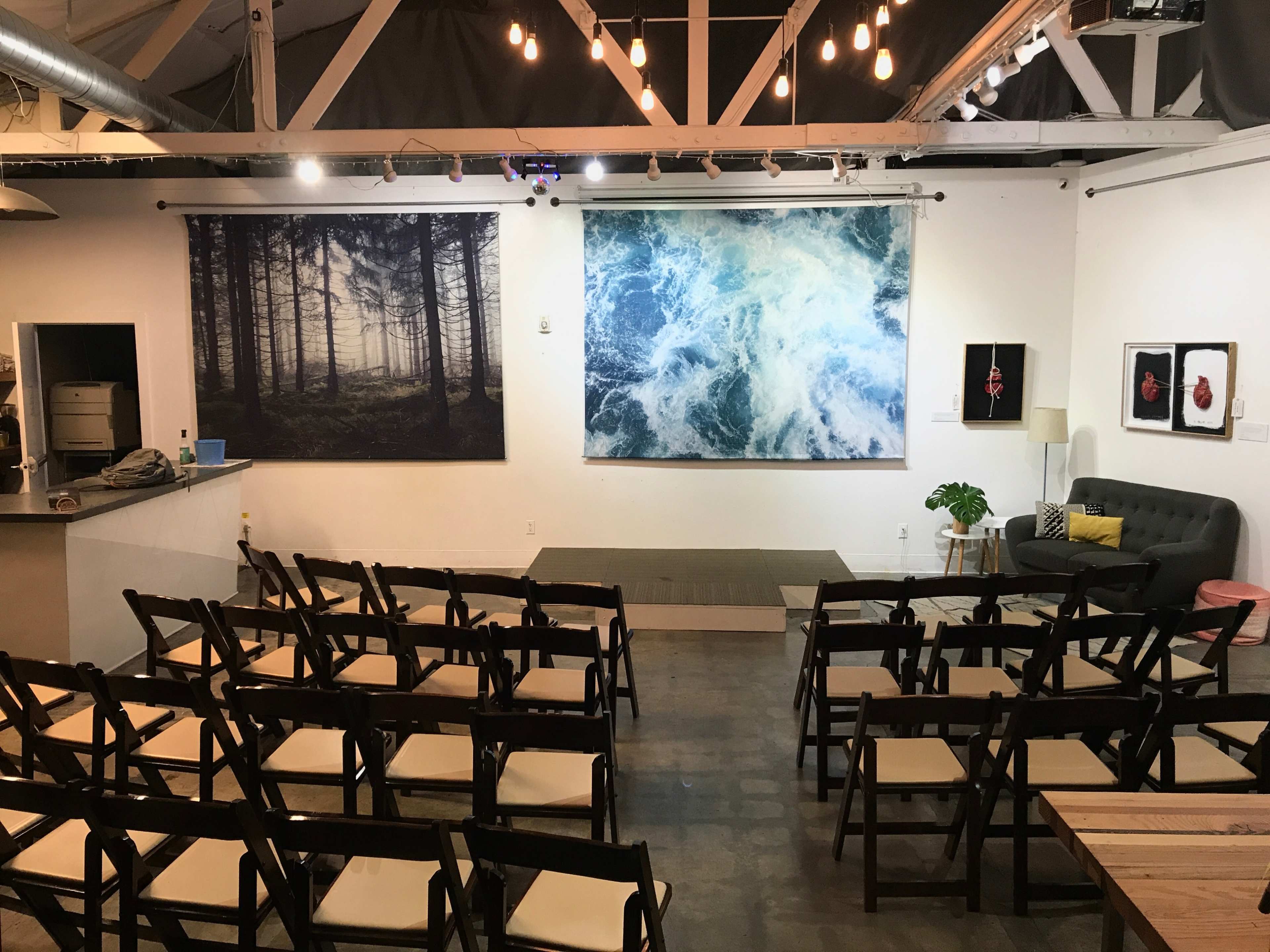 The image shows an art space with two large wall-mounted photographs and rows of empty folding chairs arranged for an event.