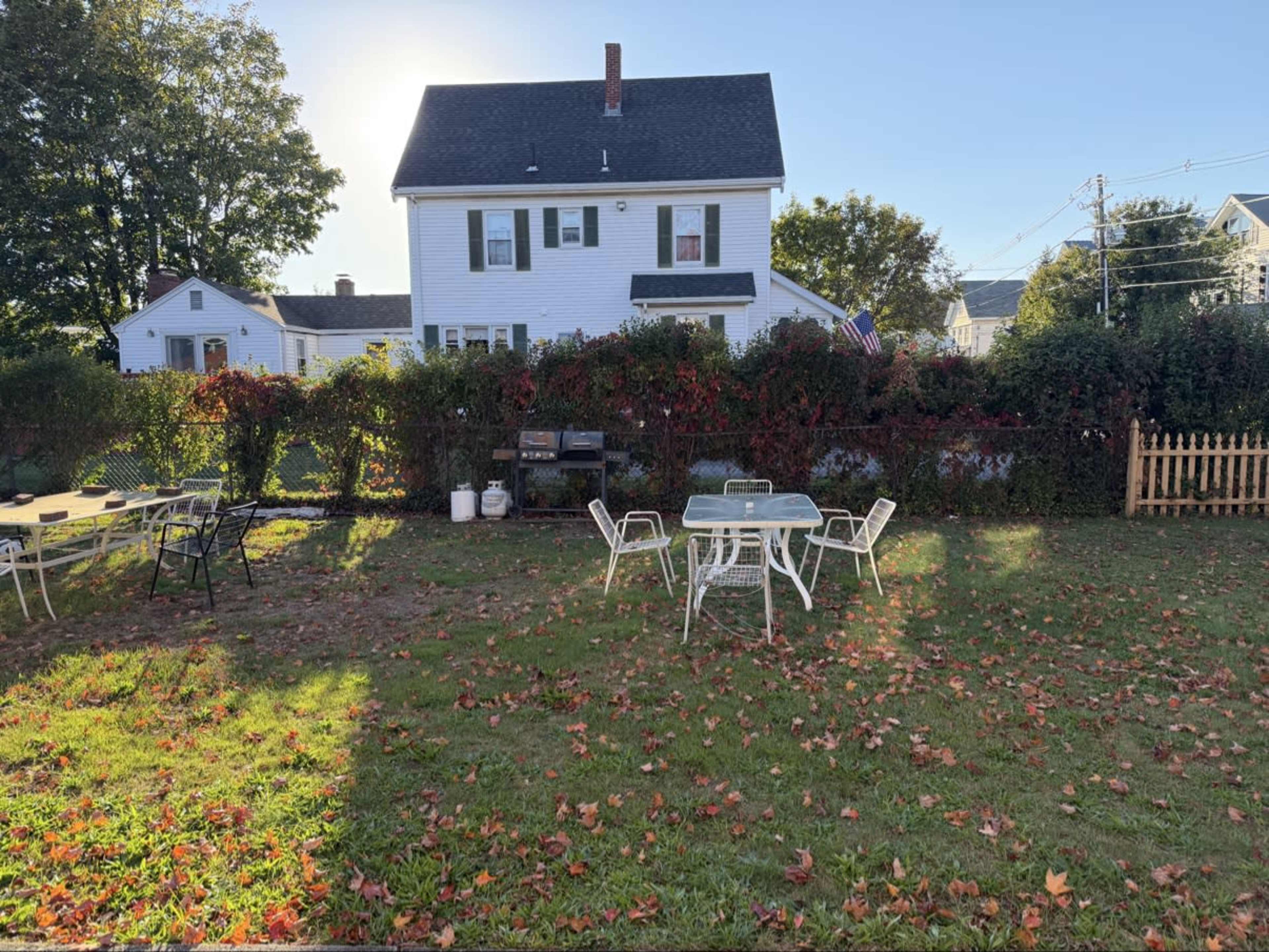 A two-story white house stands behind a yard scattered with fallen leaves and a table set with chairs.