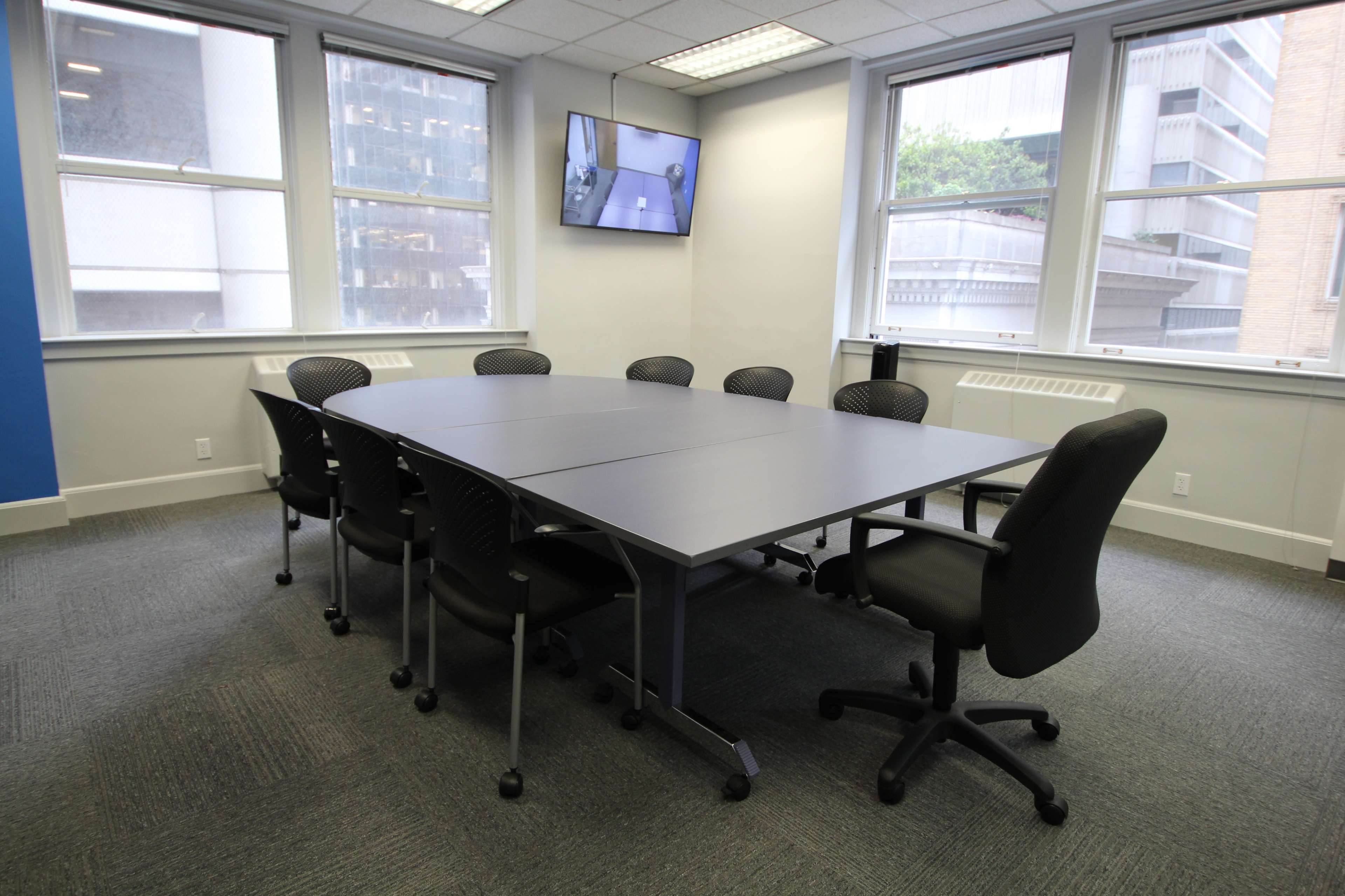 A conference room features a large rectangular table surrounded by eight black rolling chairs, with large windows and a wall-mounted television.
