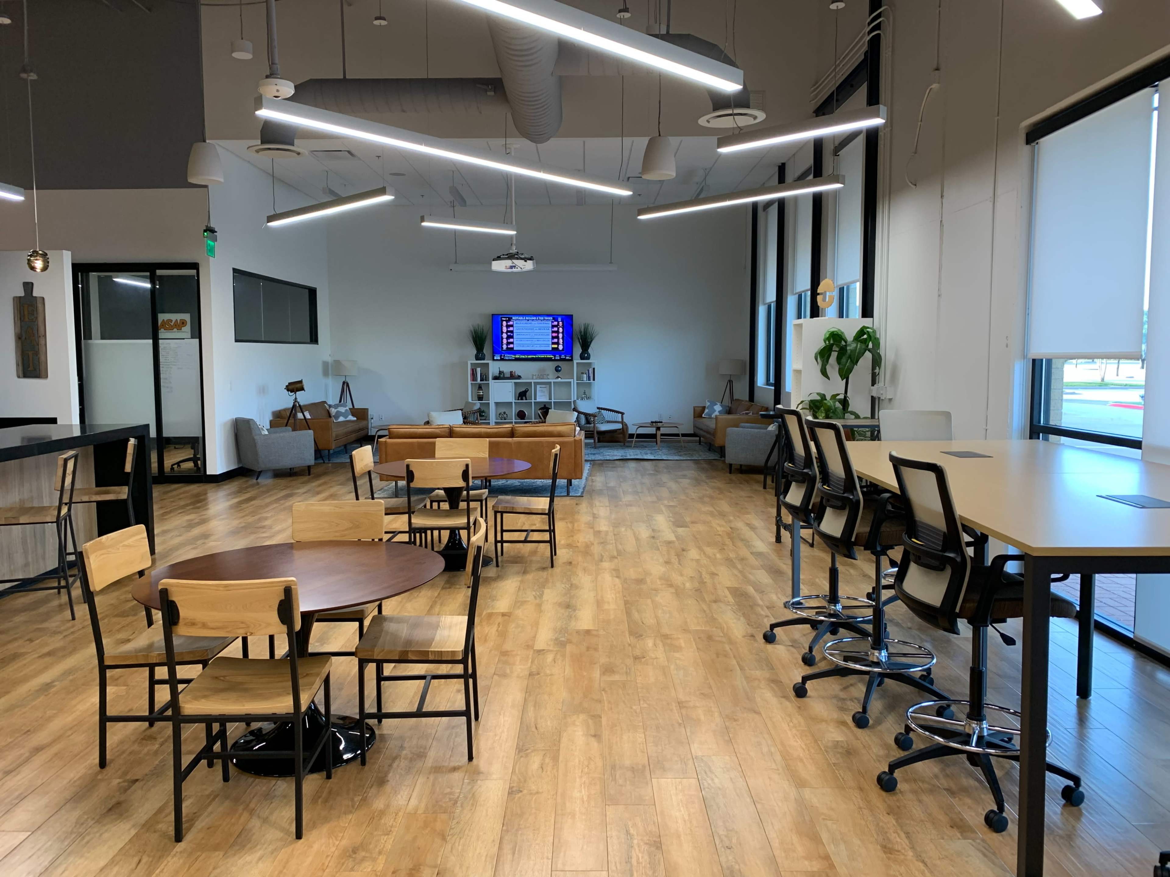A modern office space featuring various seating arrangements, workstations, and a large screen mounted on the wall.