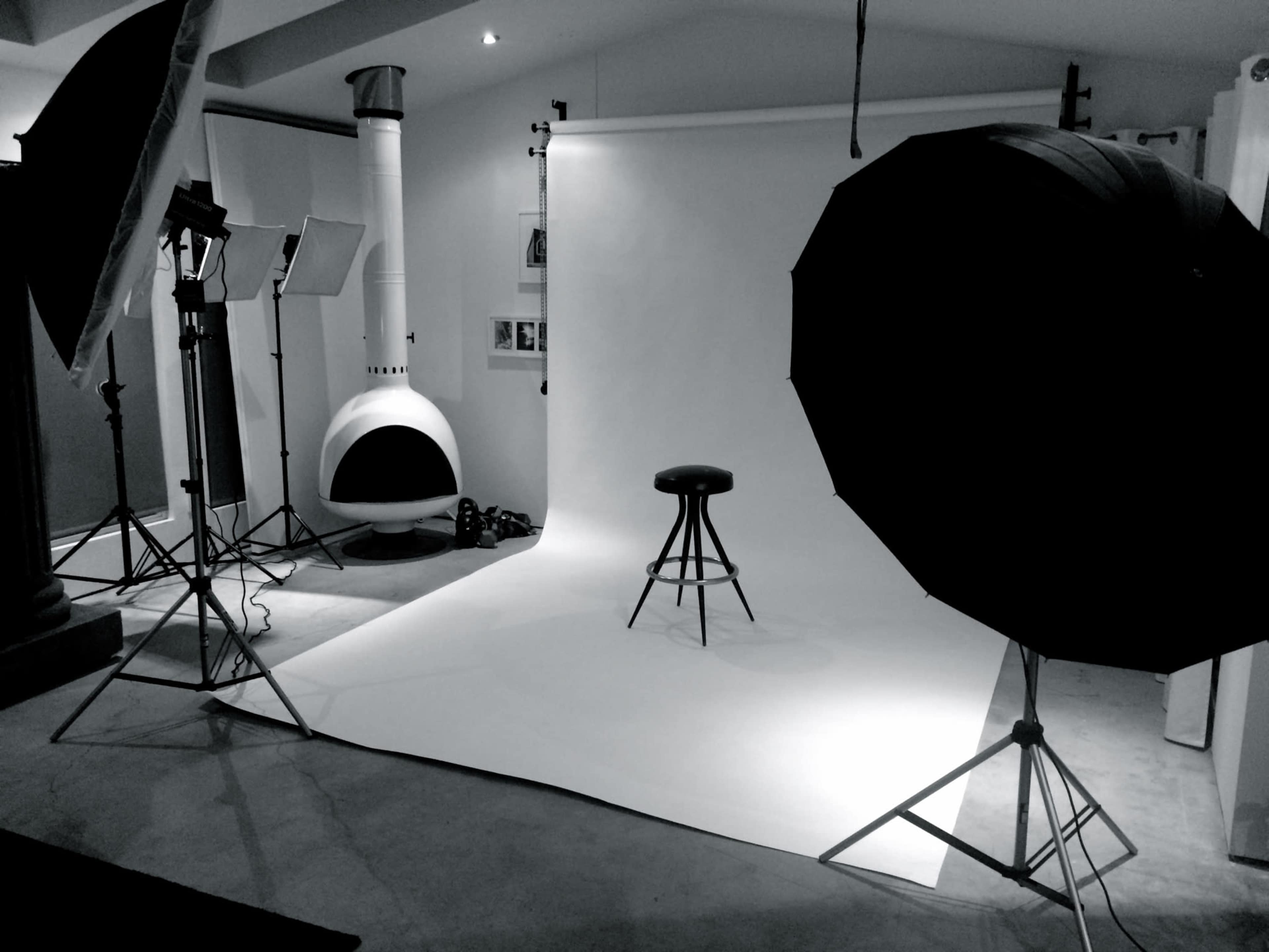 The image shows a photography studio with a plain backdrop, a stool positioned in the center, and several lighting equipment setups around the area.