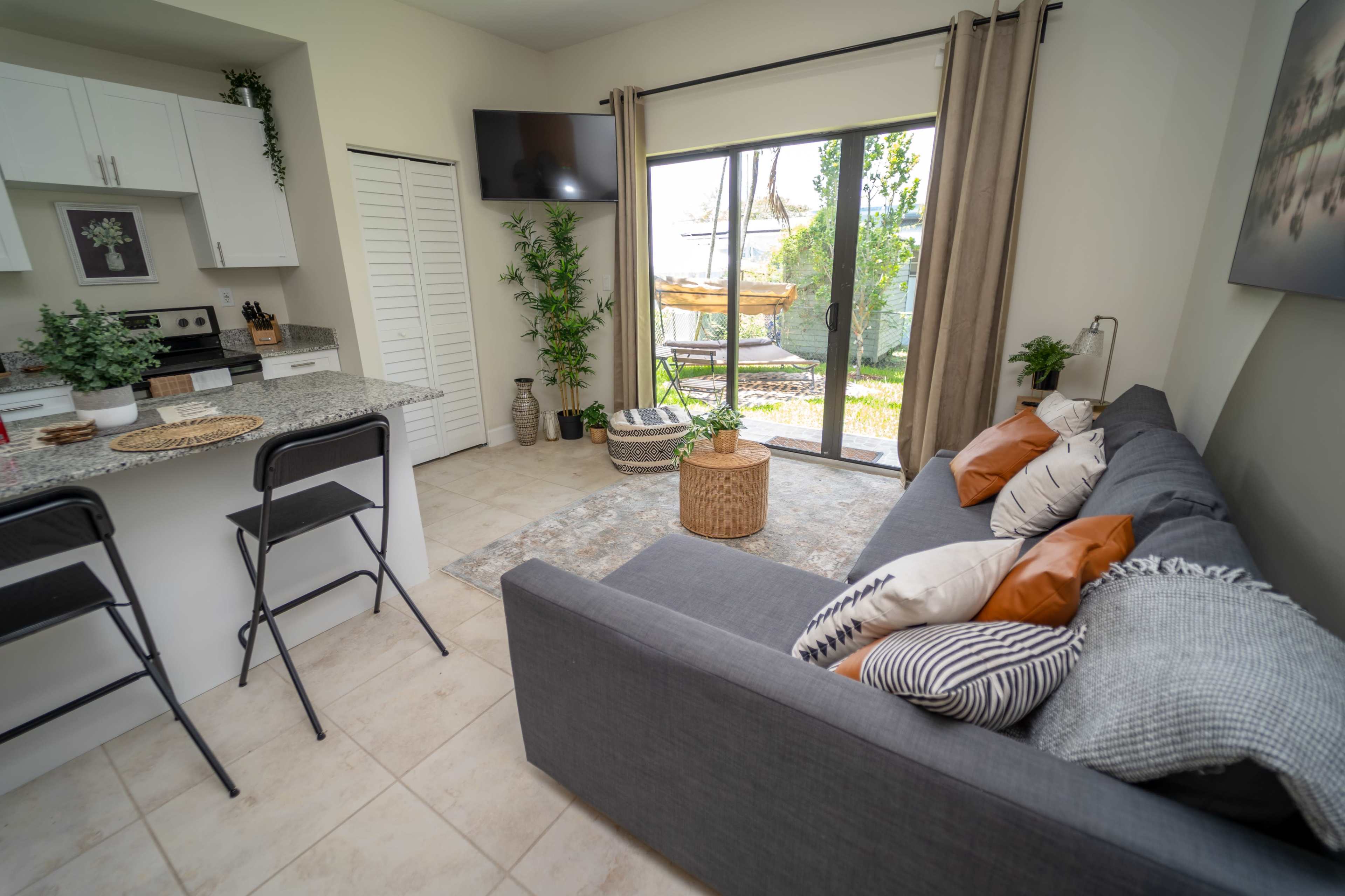 The image shows a modern living space with a gray sofa, a decorative area rug, a small dining area with stools, and a glass door leading to an outdoor area.