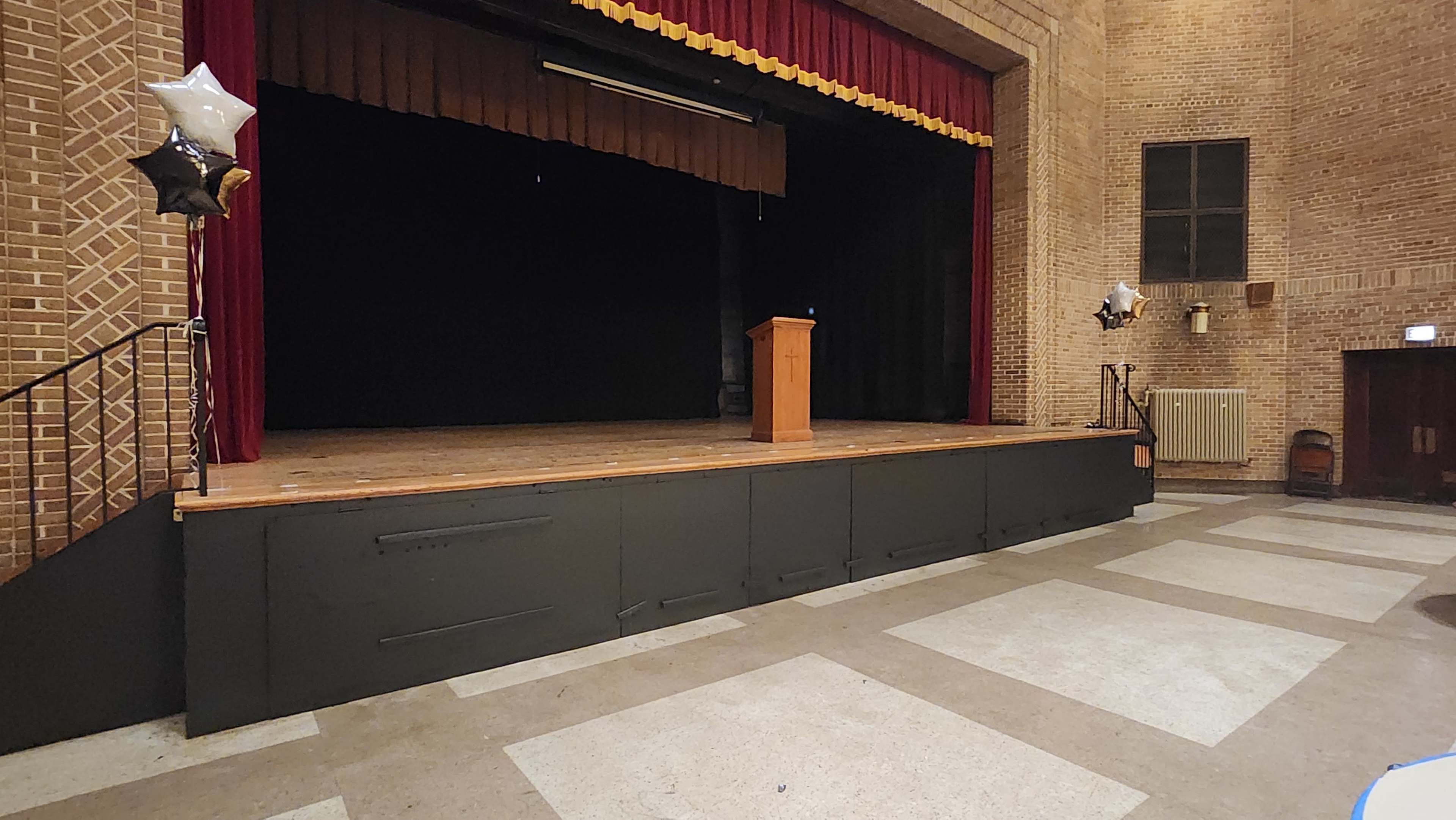 The image shows a stage with a podium in a brick auditorium, featuring a dark curtain and decorations of balloons nearby.