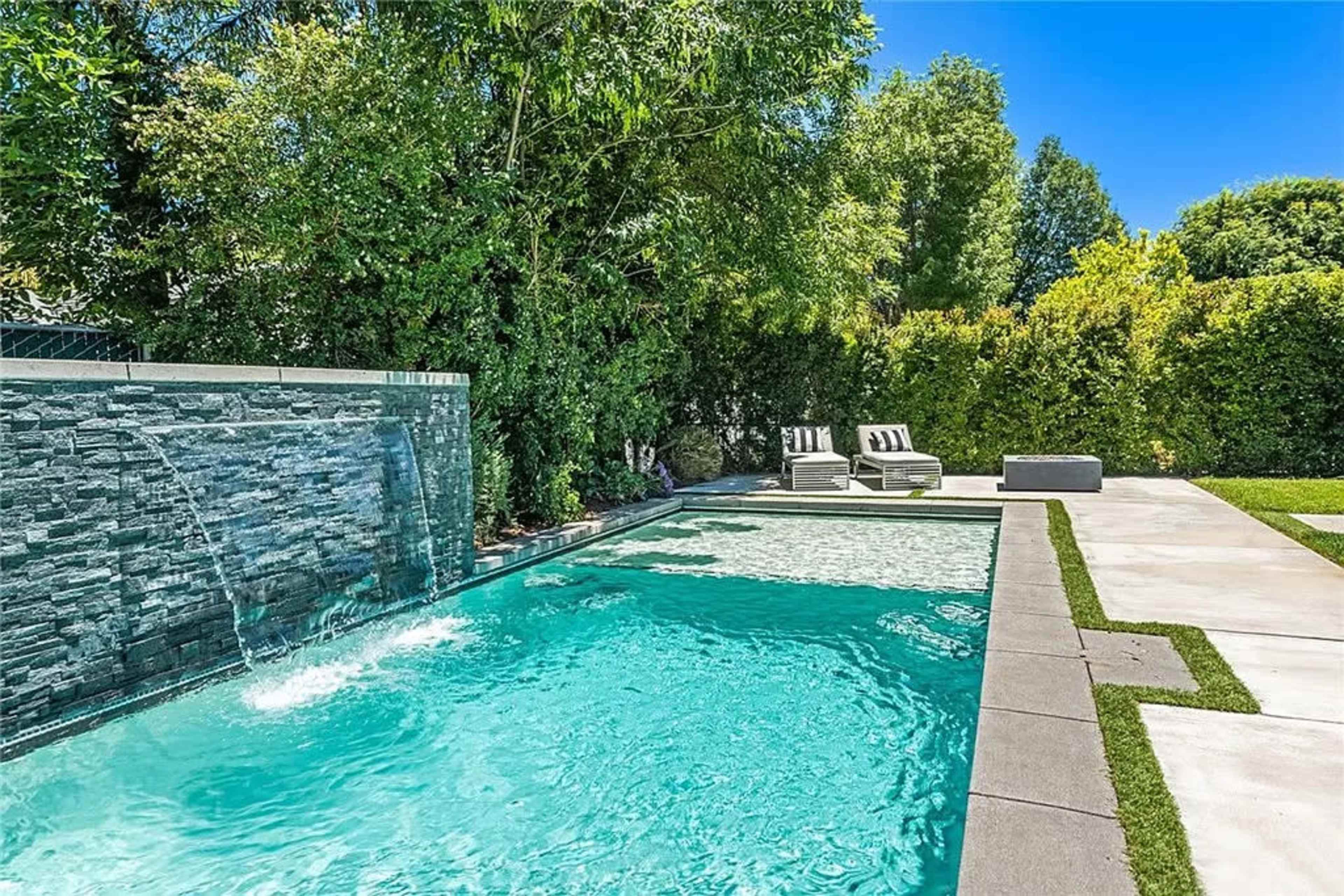 A backyard pool featuring a stone waterfall and two lounge chairs beside a landscaped area with greenery.