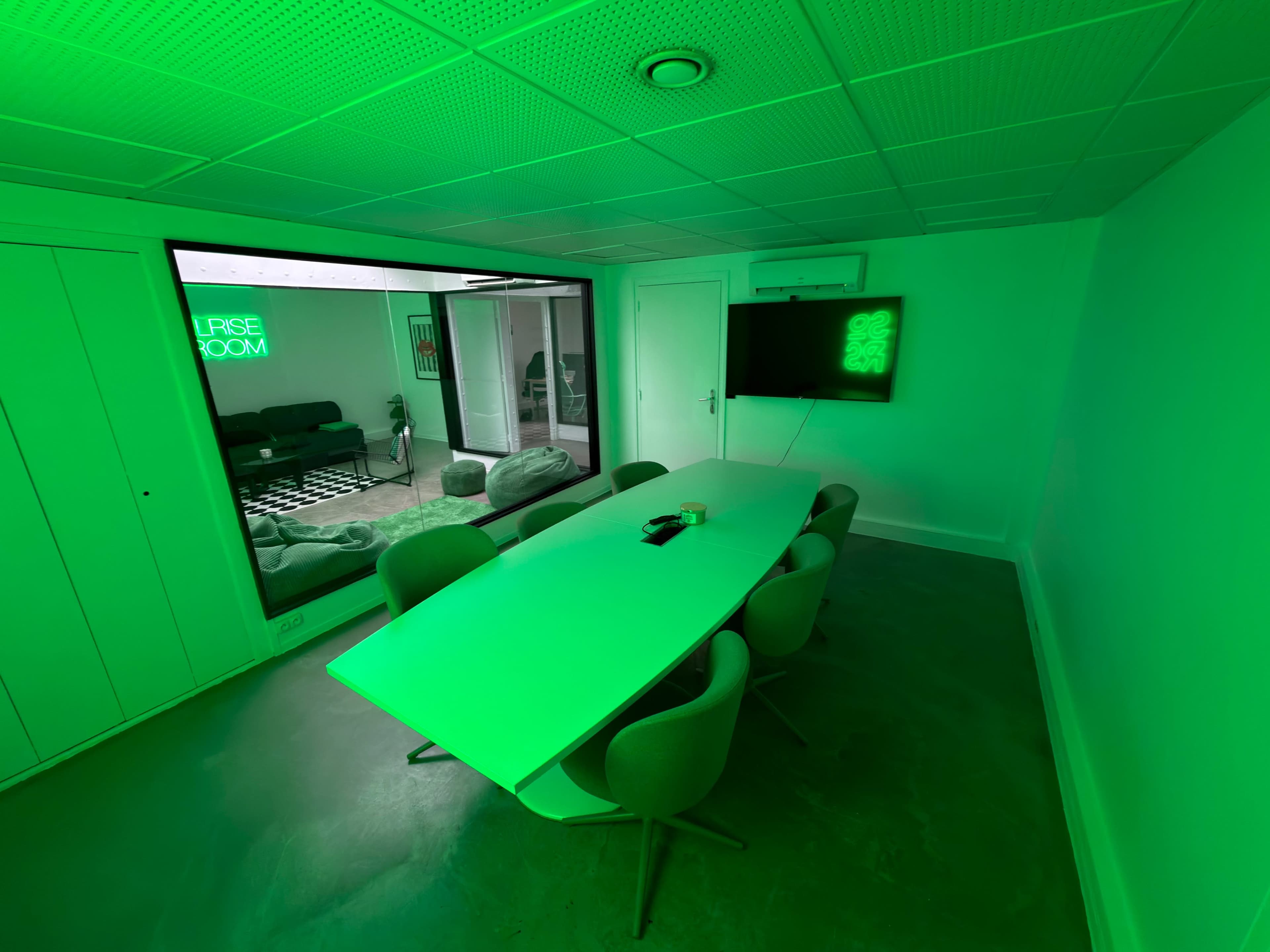 A modern meeting room features a long white table surrounded by gray chairs, illuminated by green lighting.