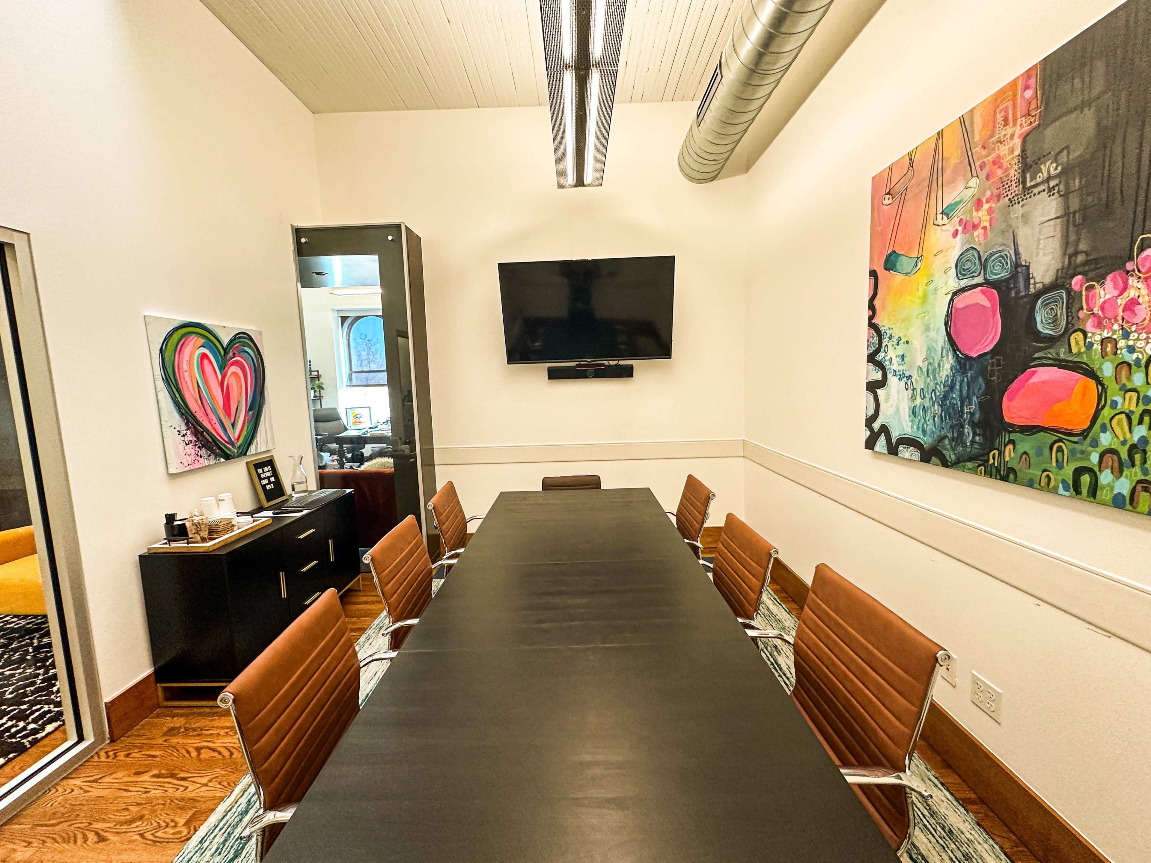 The image shows a meeting room with a long black conference table, eight brown leather chairs, and colorful abstract paintings on the walls.