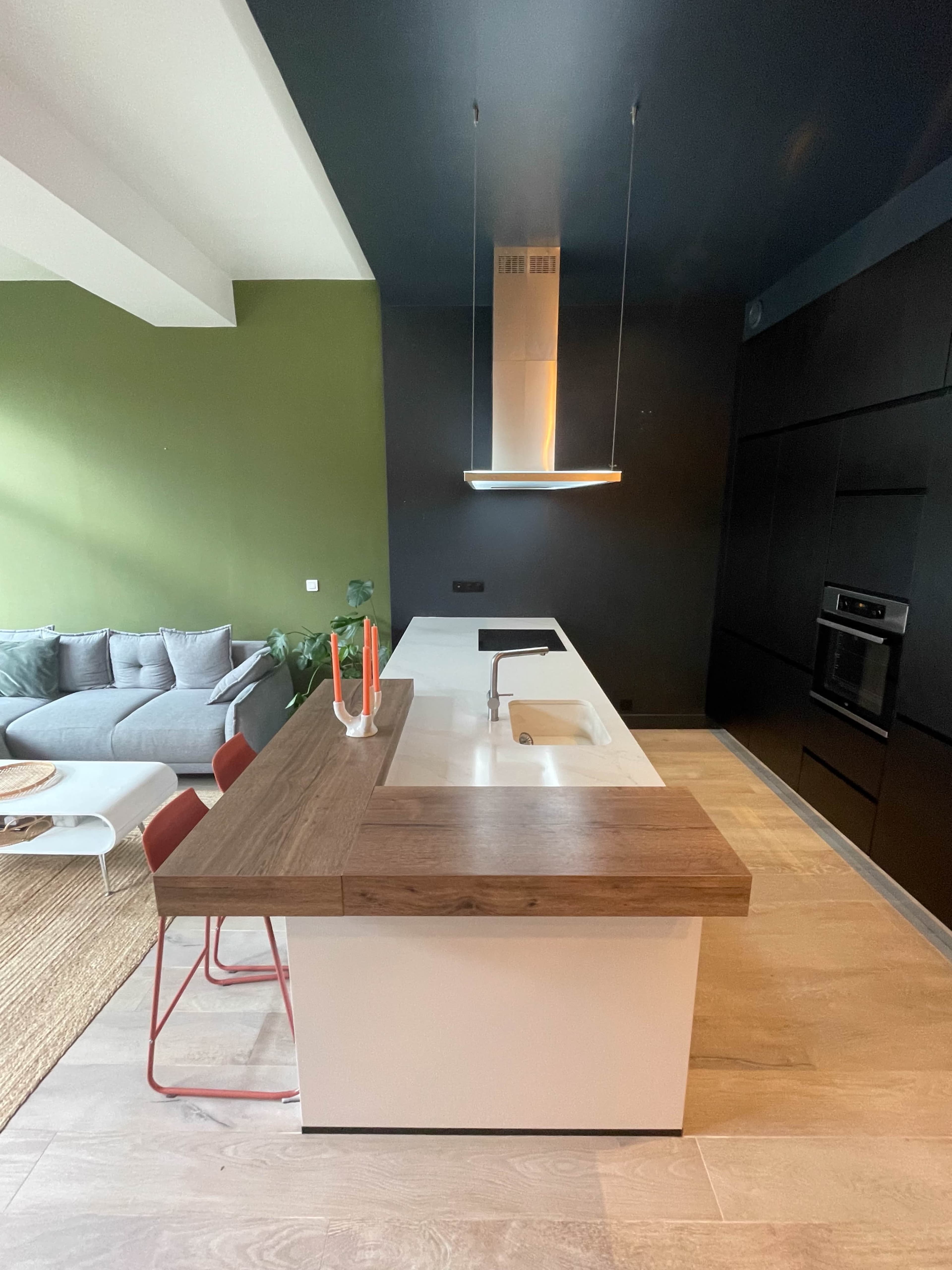 The image shows a modern kitchen featuring a central island with a wooden countertop, a sink, and a minimalist design, adjacent to a living area with a gray sofa and a green accent wall.