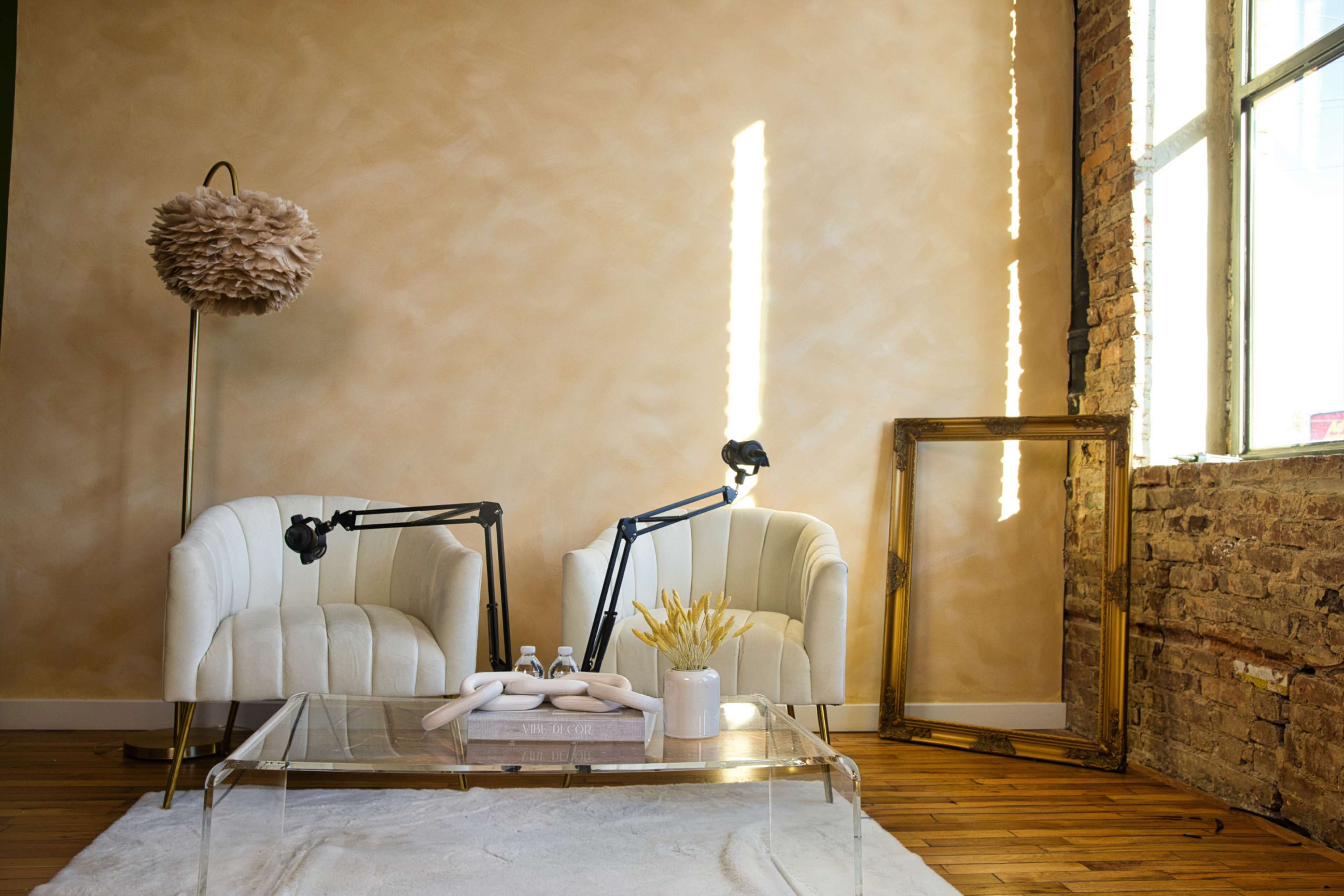 The image shows a stylish sitting area with two white chairs, a glass coffee table, and a soft rug, set against a wall of warm-toned plaster and a large window.