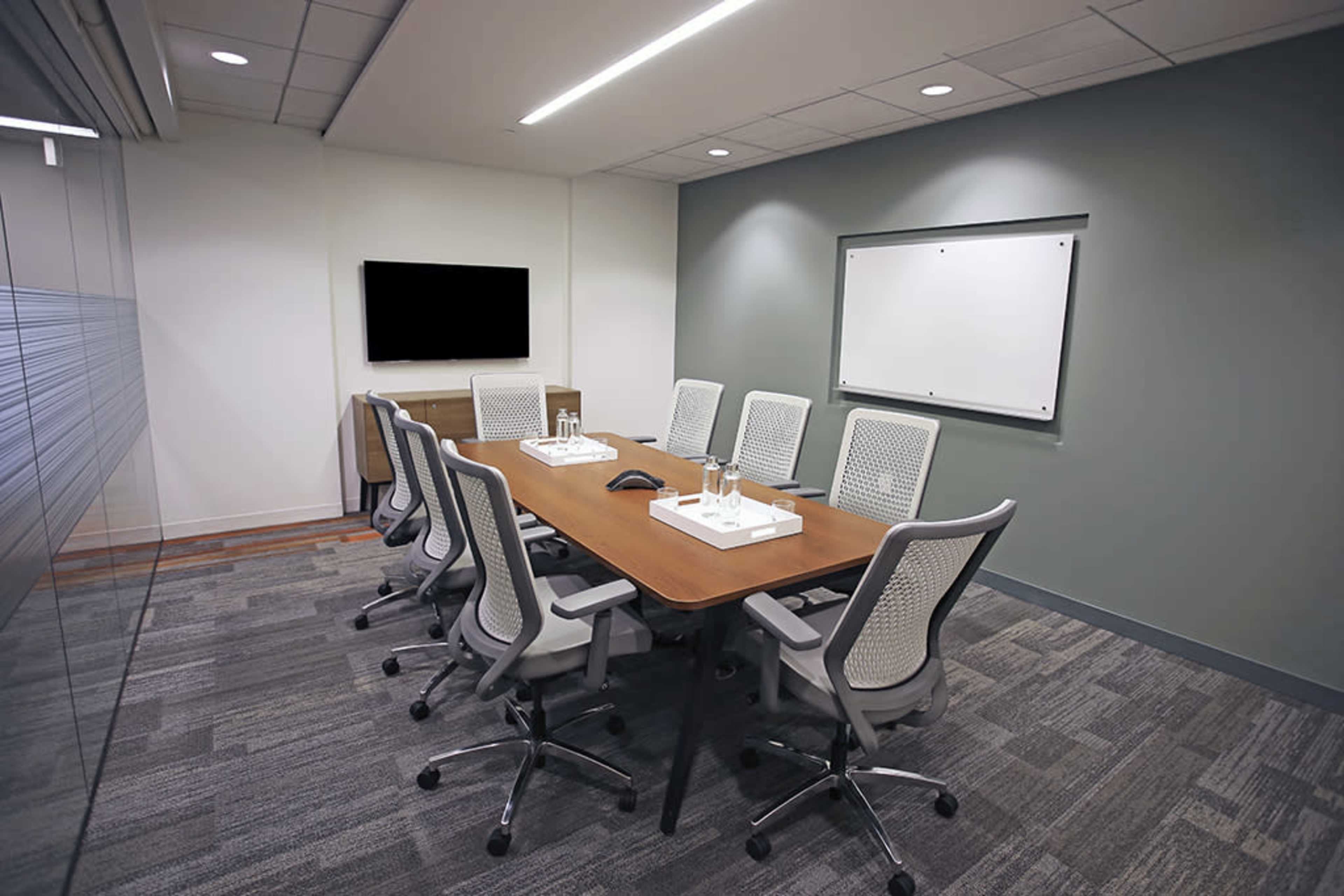 A modern conference room features a long table surrounded by ergonomic chairs, with a television and whiteboard on the walls.