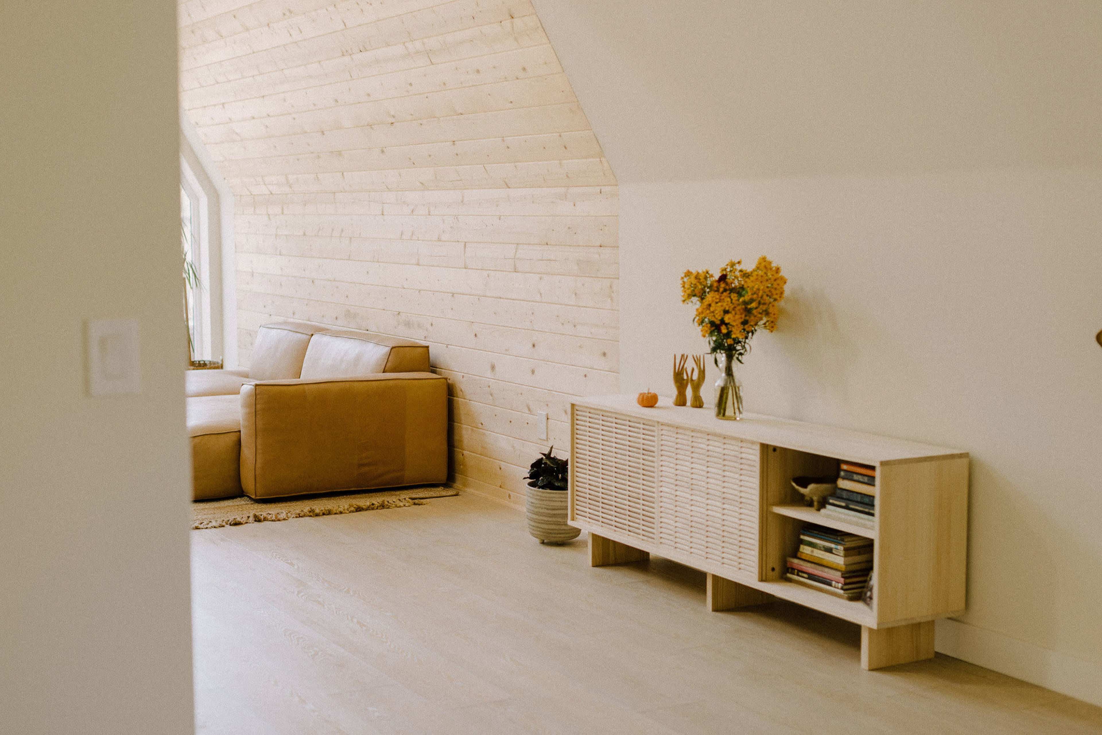 A light-colored living room features a wooden wall, a tan couch, a sideboard with books and decorative items, and a bunch of flowers on top.