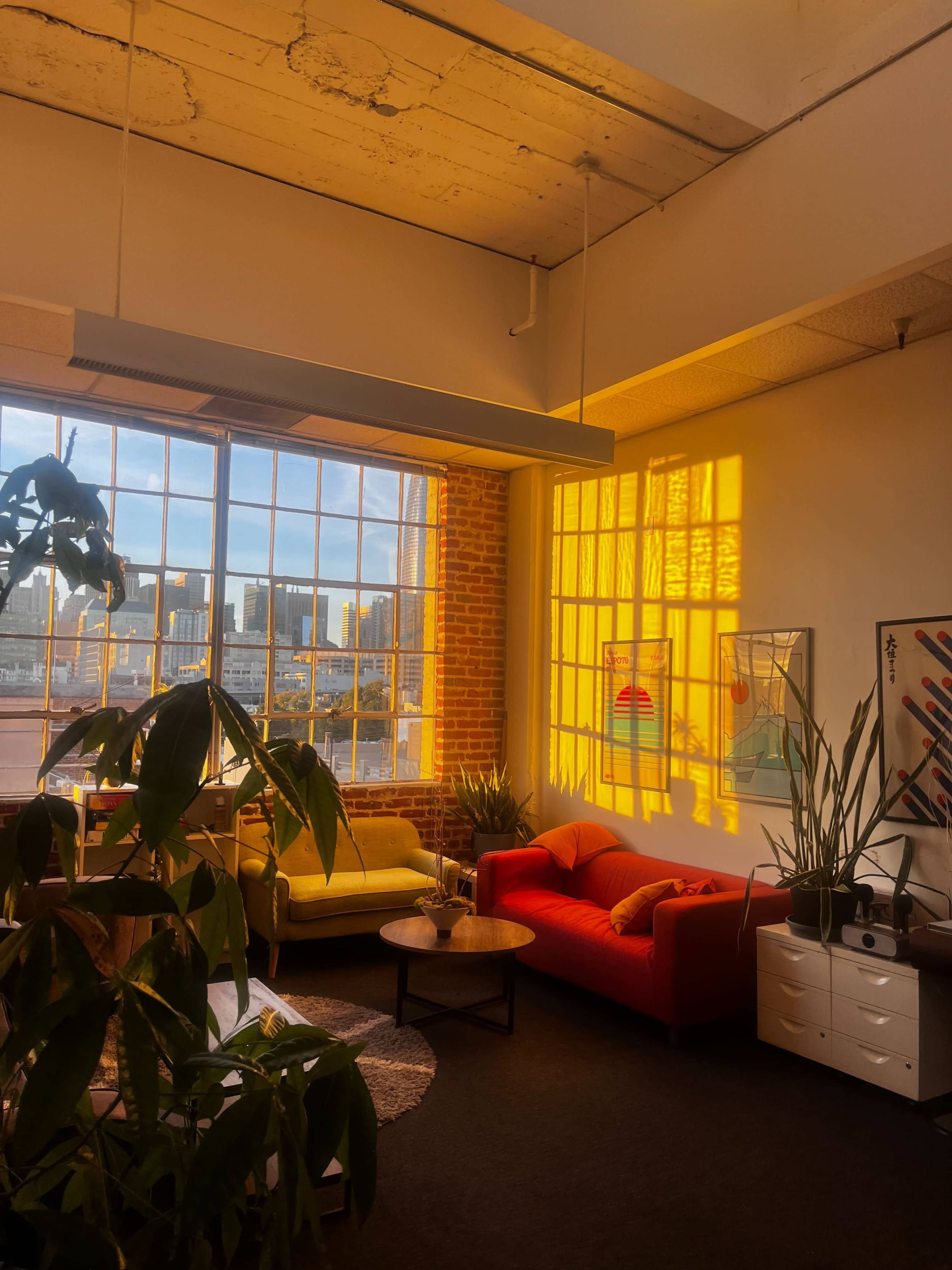 A spacious room features large windows that illuminate a yellow sofa and a red couch, with plants and artwork enhancing the decor.