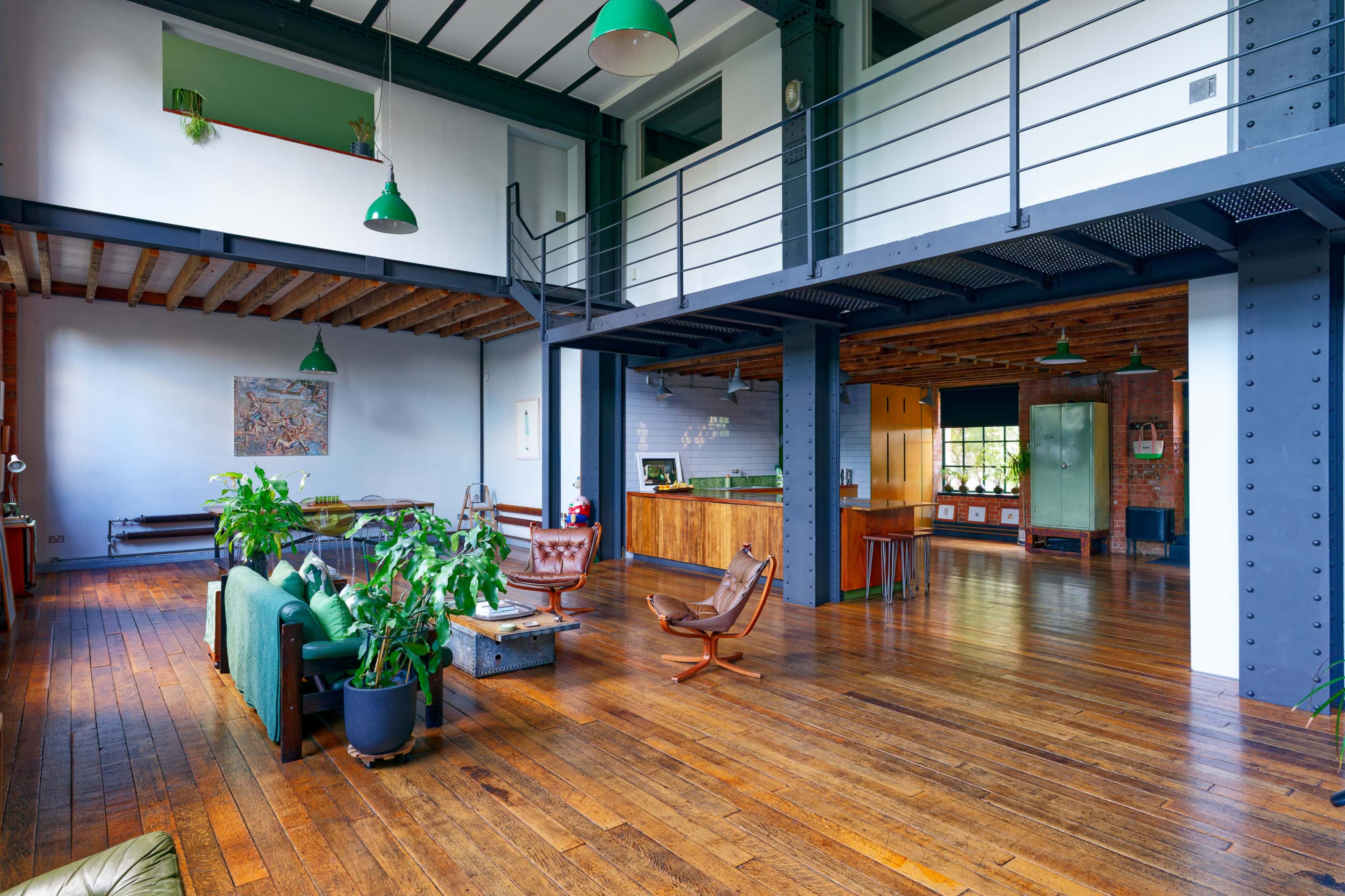 The image shows an open-concept living space with wooden flooring, industrial-style elements, and greenery integrated throughout the design.