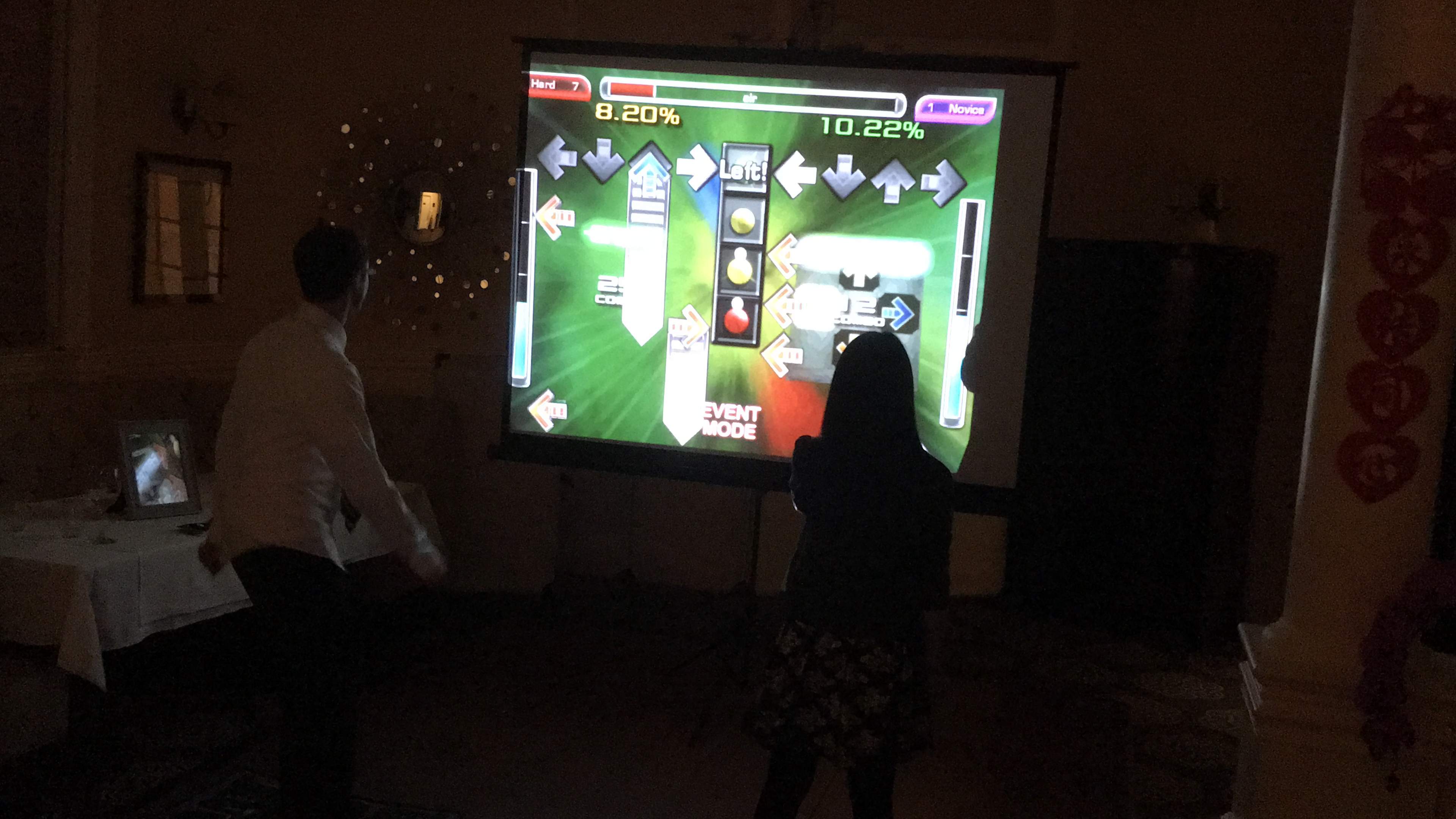Two people are playing a rhythm game on a large projected screen in a dimly lit room.
