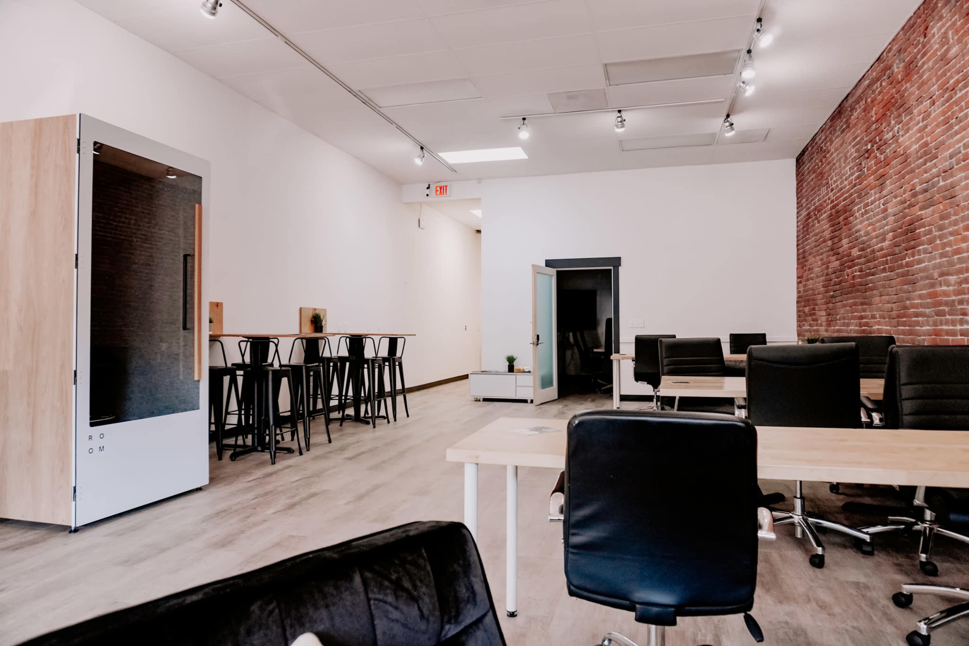 A modern office interior featuring wooden desks, black chairs, tall bar-style seating, and exposed brick walls.