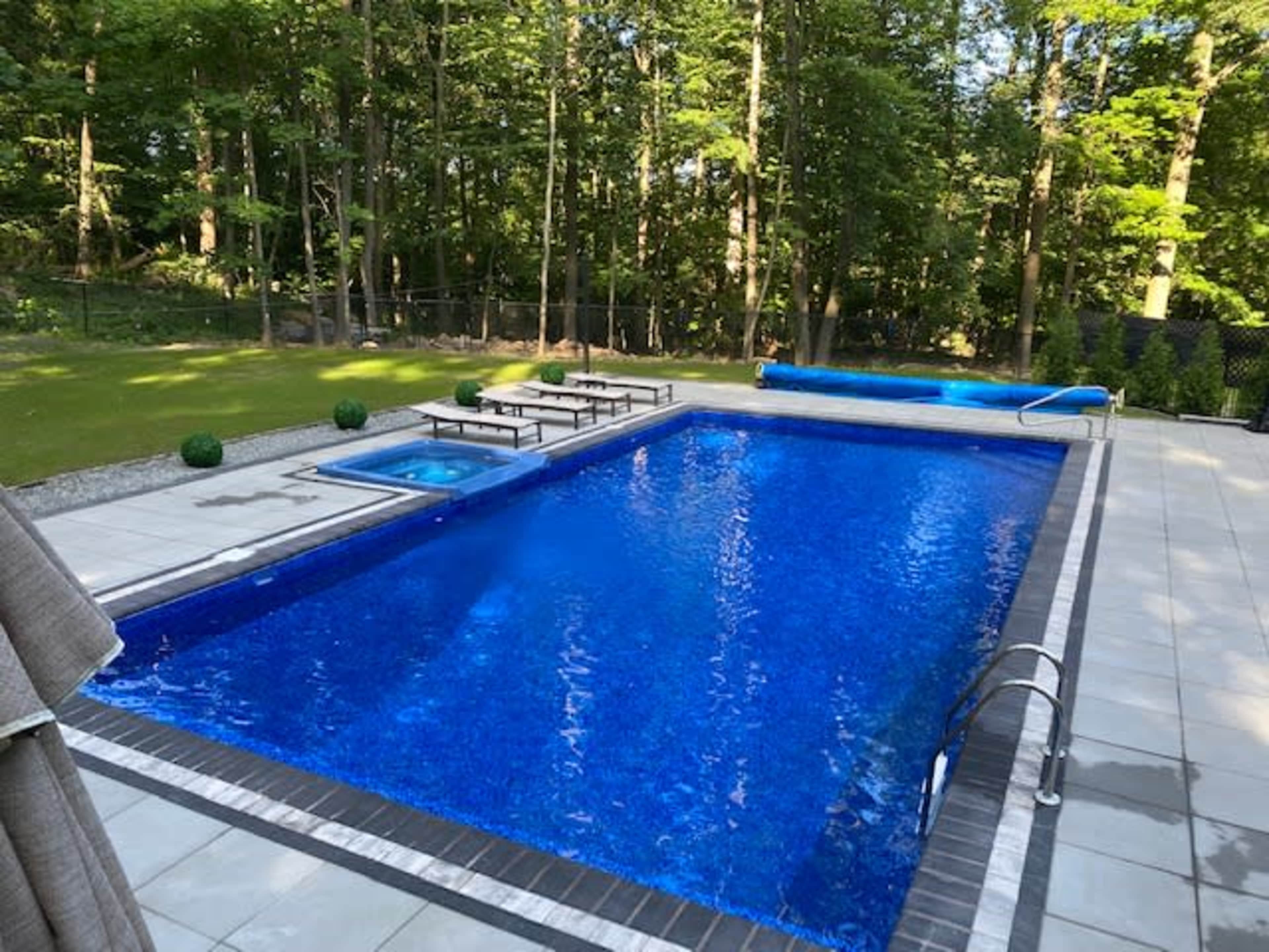 A large, blue swimming pool surrounded by stone decking, with lounge chairs and a smaller spa area nearby, set in a wooded backyard.