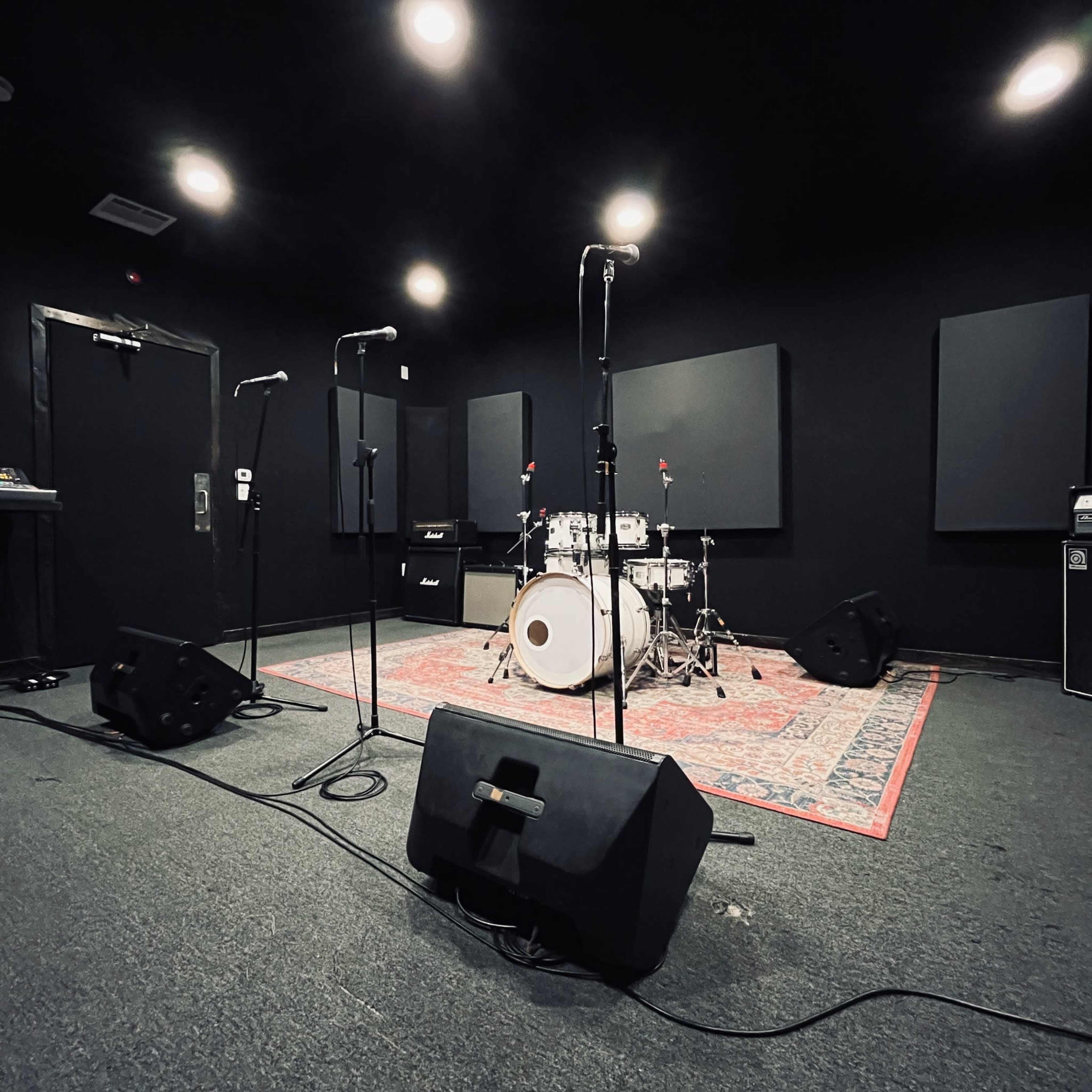 The image shows a rehearsal studio with drum set, microphones on stands, and amplifiers placed against black walls and a patterned rug on the floor.