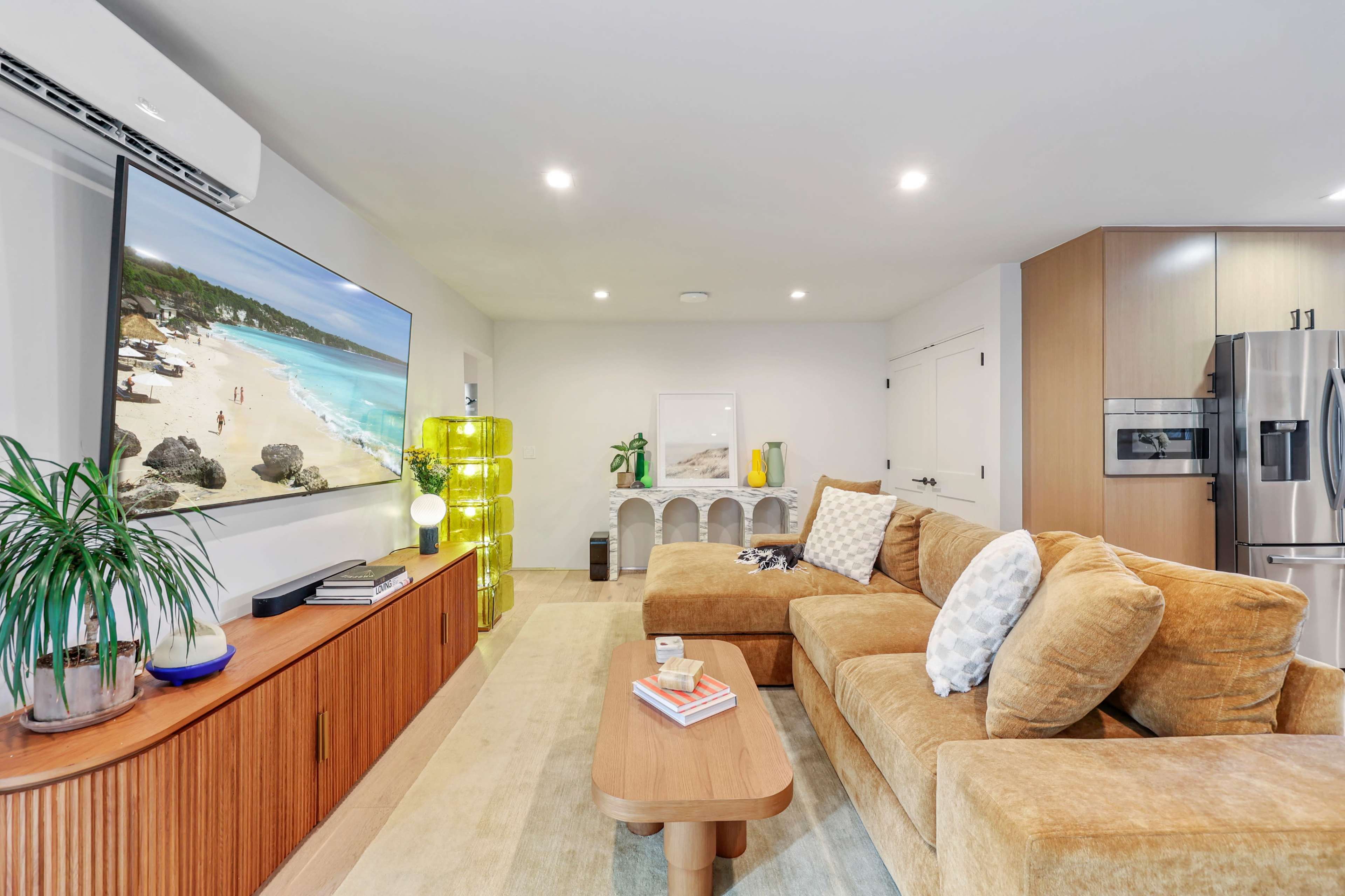 A modern living room features a large flat-screen TV, a brown sectional couch, a wooden sideboard, and a bright yellow lamp, with a large window illuminating the space.