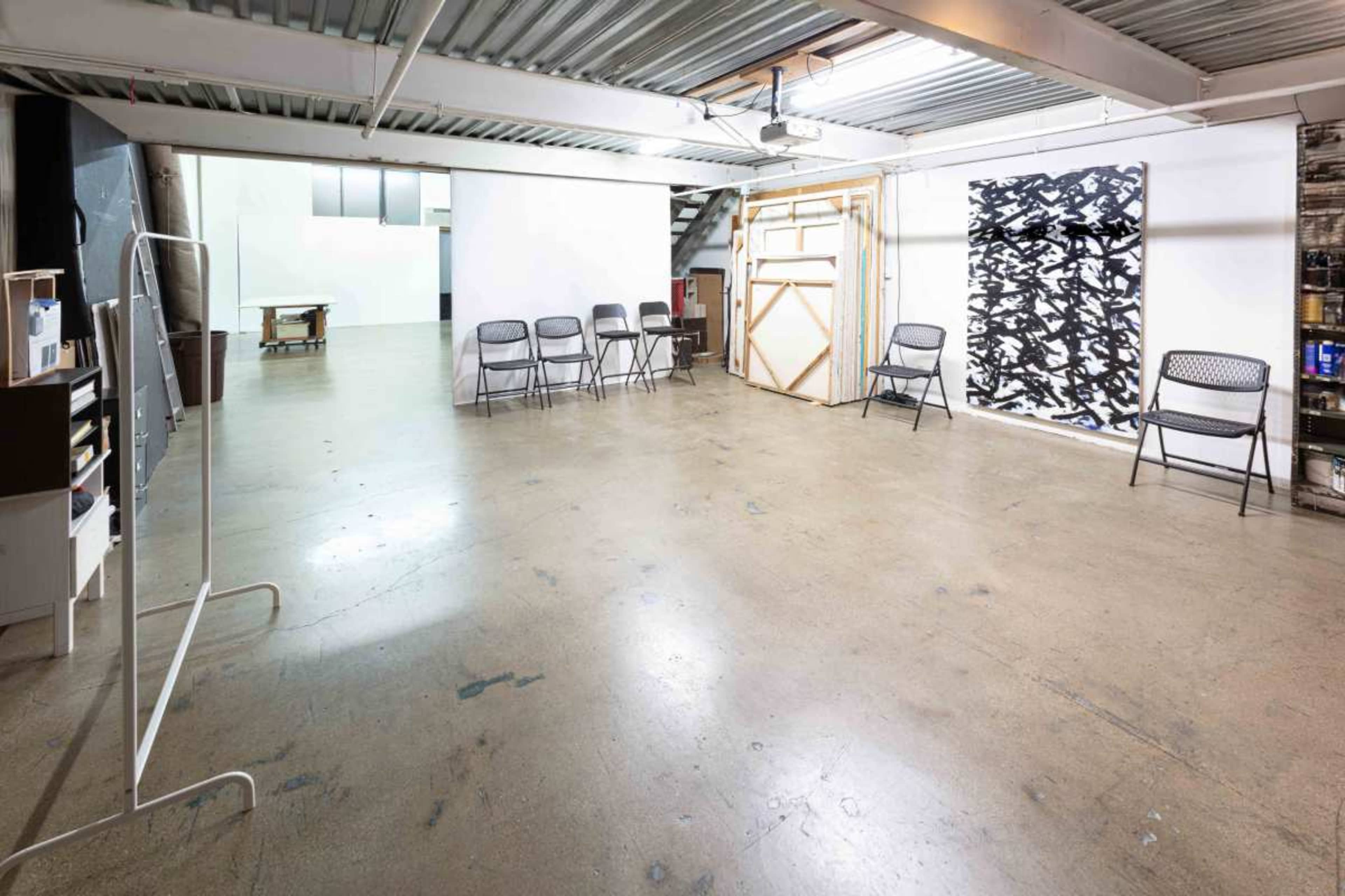 The image shows a spacious art studio with bare concrete flooring, several folding chairs arranged in a line, and large blank walls, one featuring a black-and-white artwork.