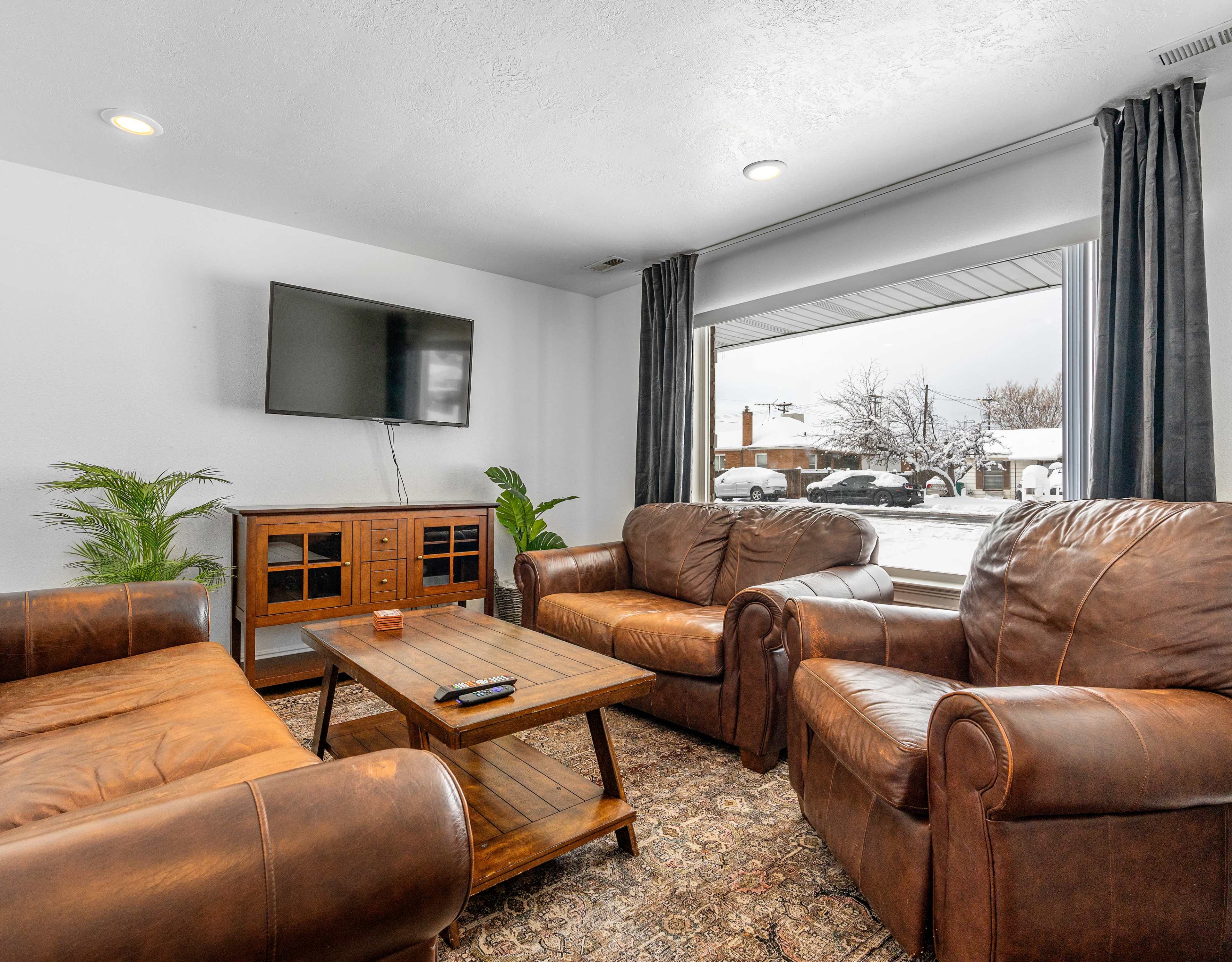 A cozy living room features two brown leather armchairs, a wooden coffee table, and a television mounted on the wall, with a view of snowy outdoor scenery through a large window.