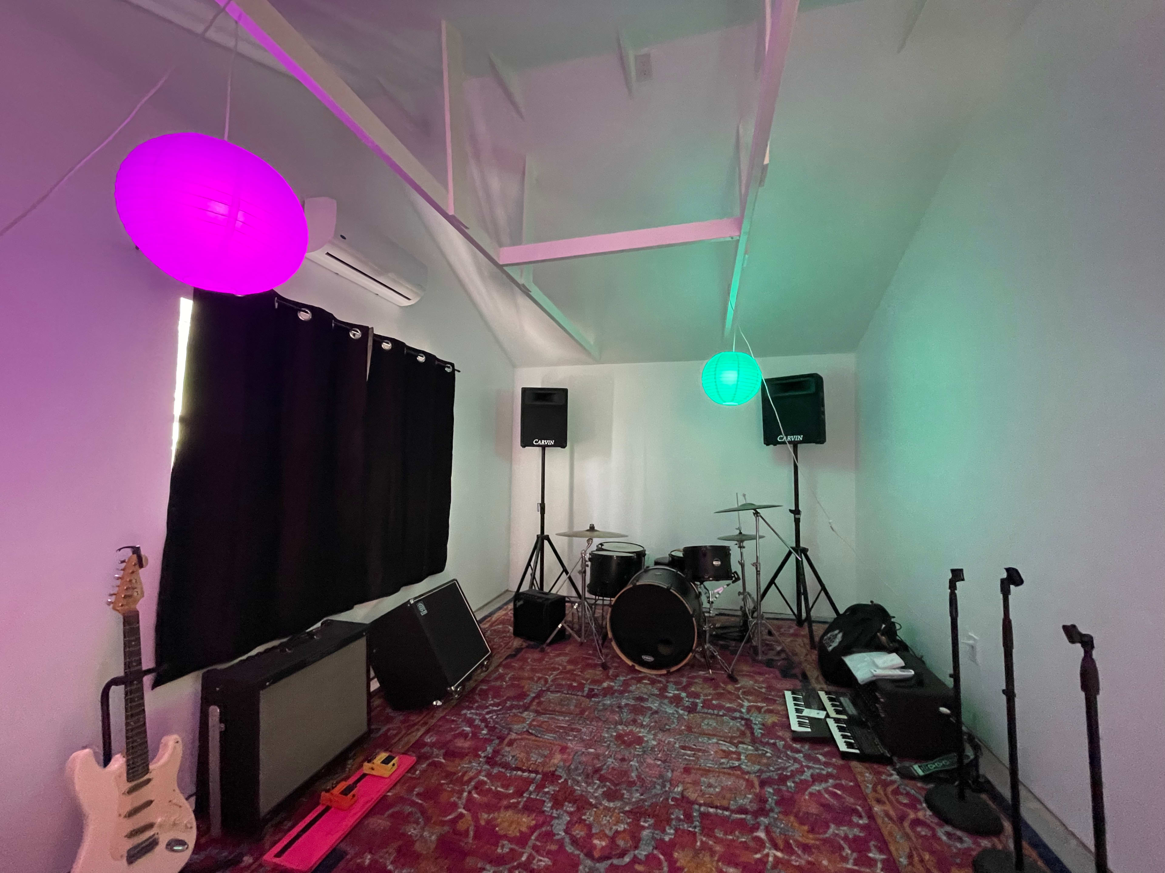 The image shows a music practice room with instruments, including a guitar and a drum set, illuminated by pink and green pendant lights.