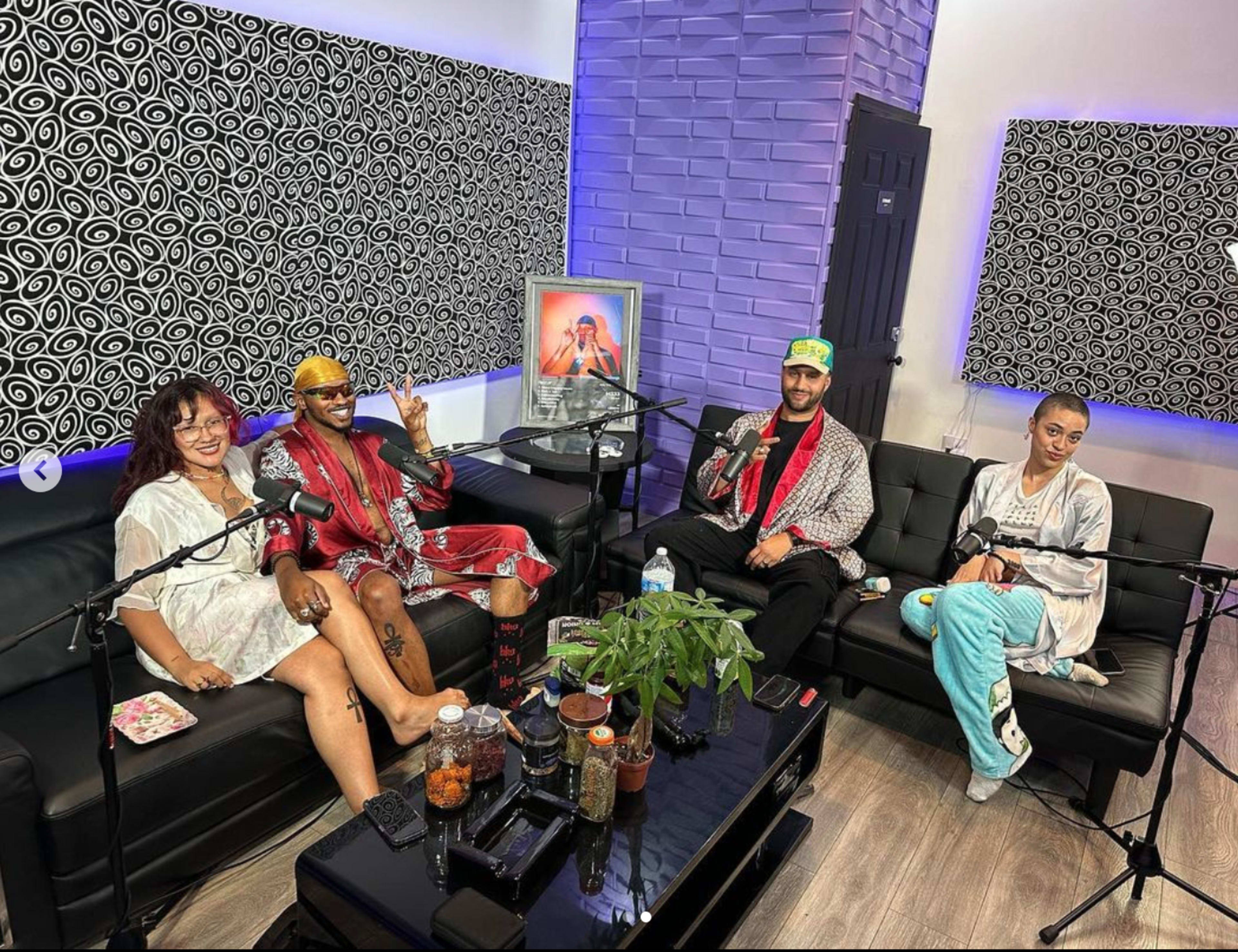 Four individuals are seated on a black couch in a brightly lit room equipped with microphones and decor featuring patterned walls.