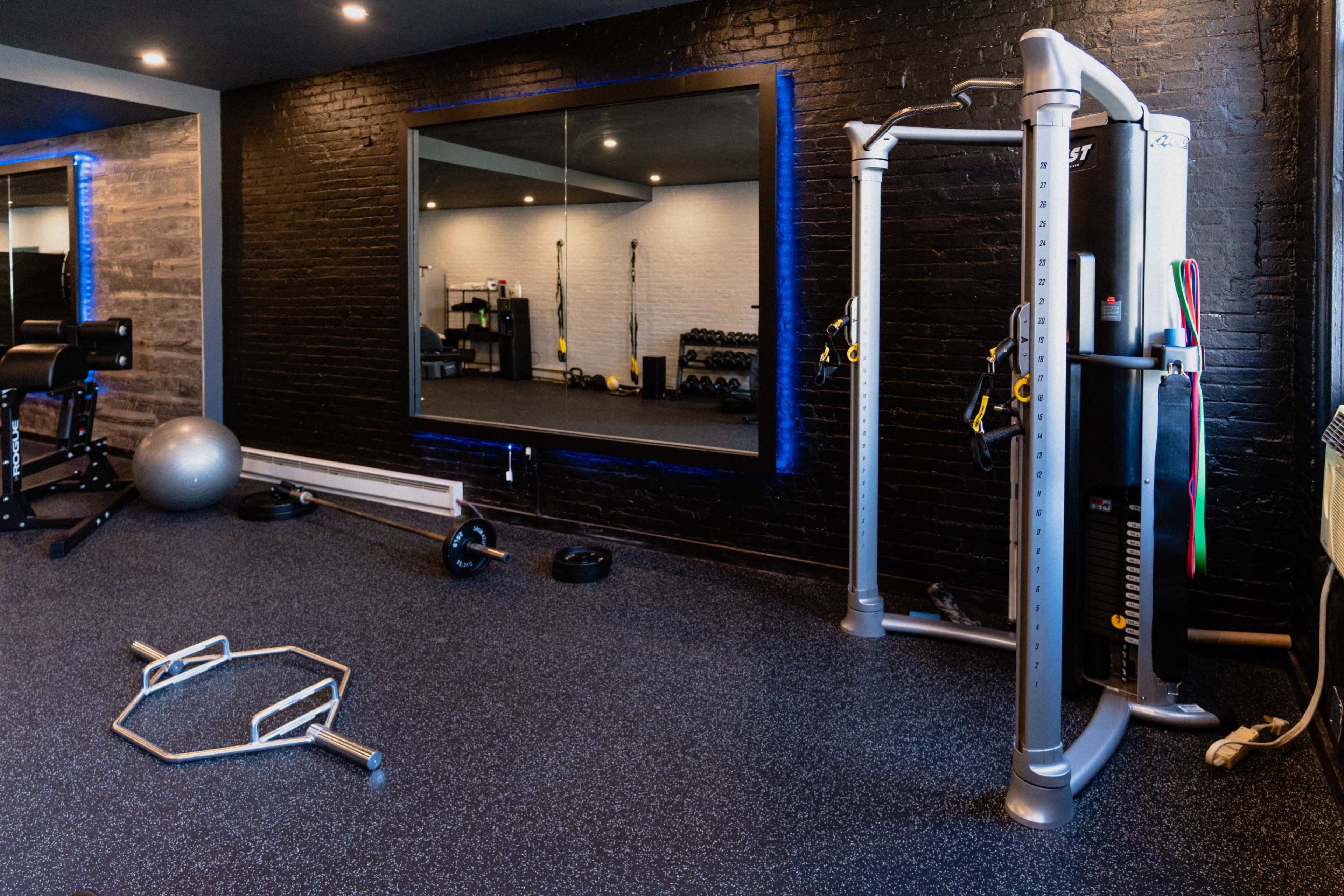 A modern gym interior featuring a cable machine, free weights, and a large mirror against black brick walls.