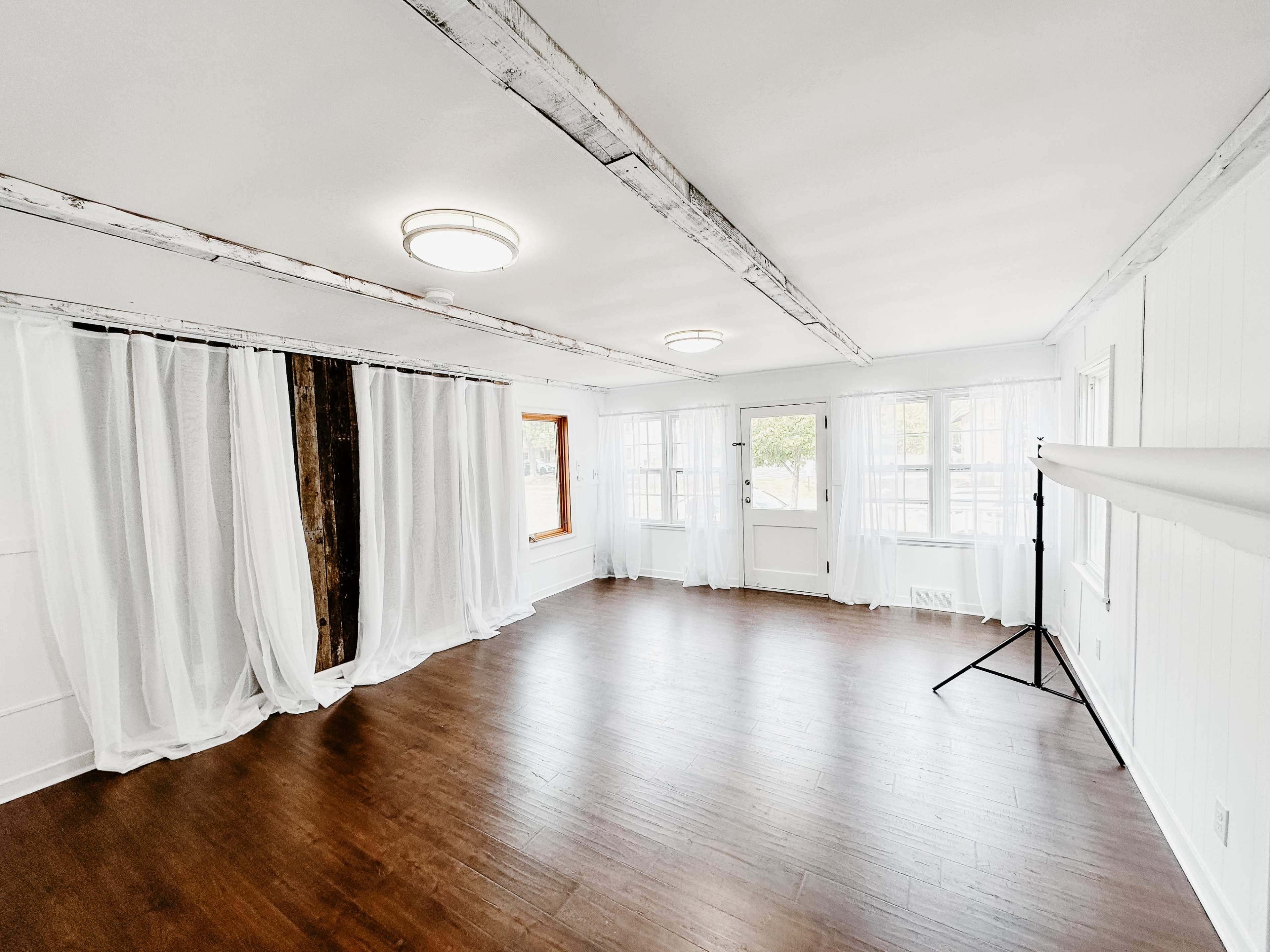 A spacious room with wooden floors, exposed beams on the ceiling, and large windows allowing natural light to enter, along with sheer curtains draped on one side.