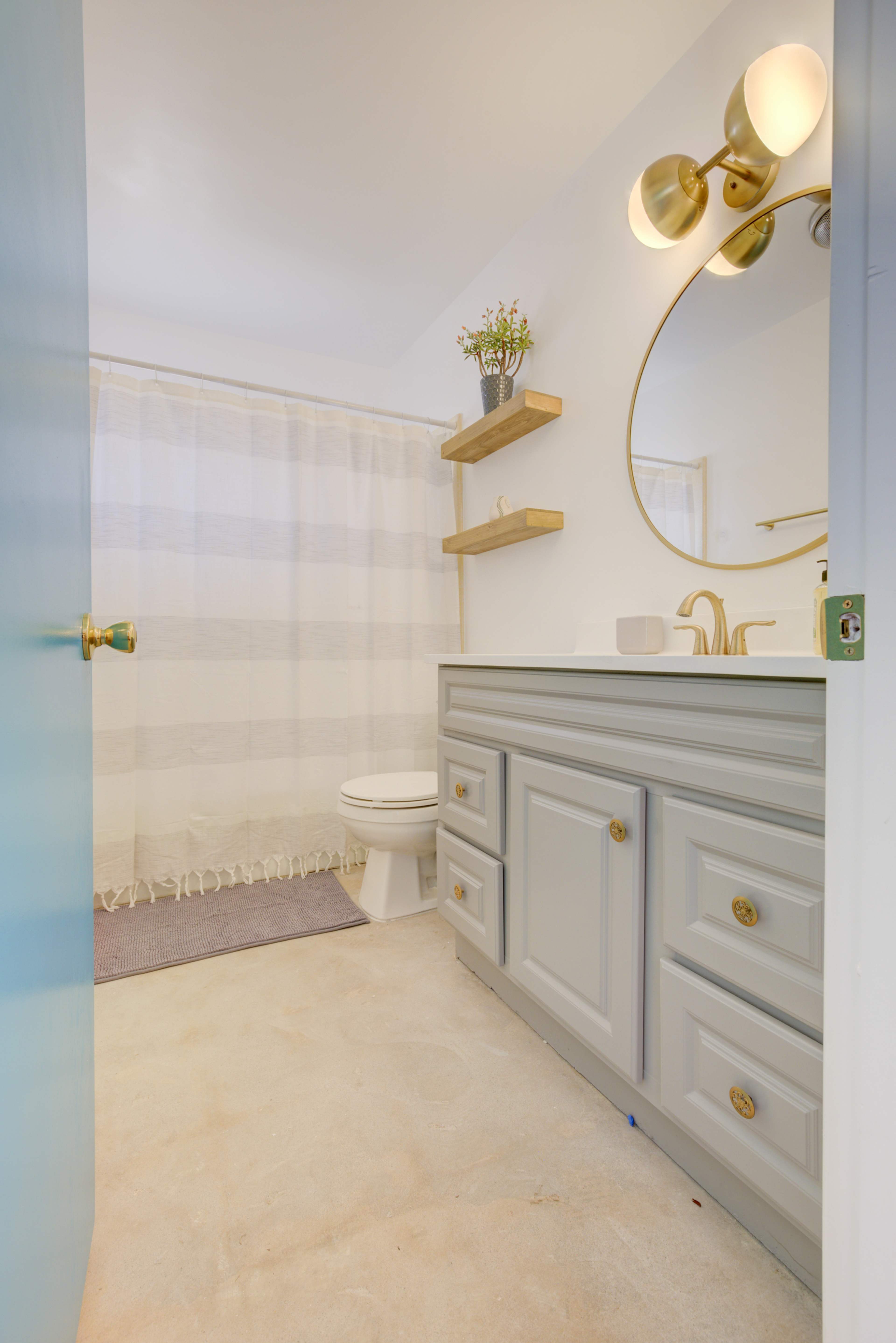 A bathroom features a light-colored vanity with gold hardware, a round mirror, and a shower curtain, with shelves above the sink.