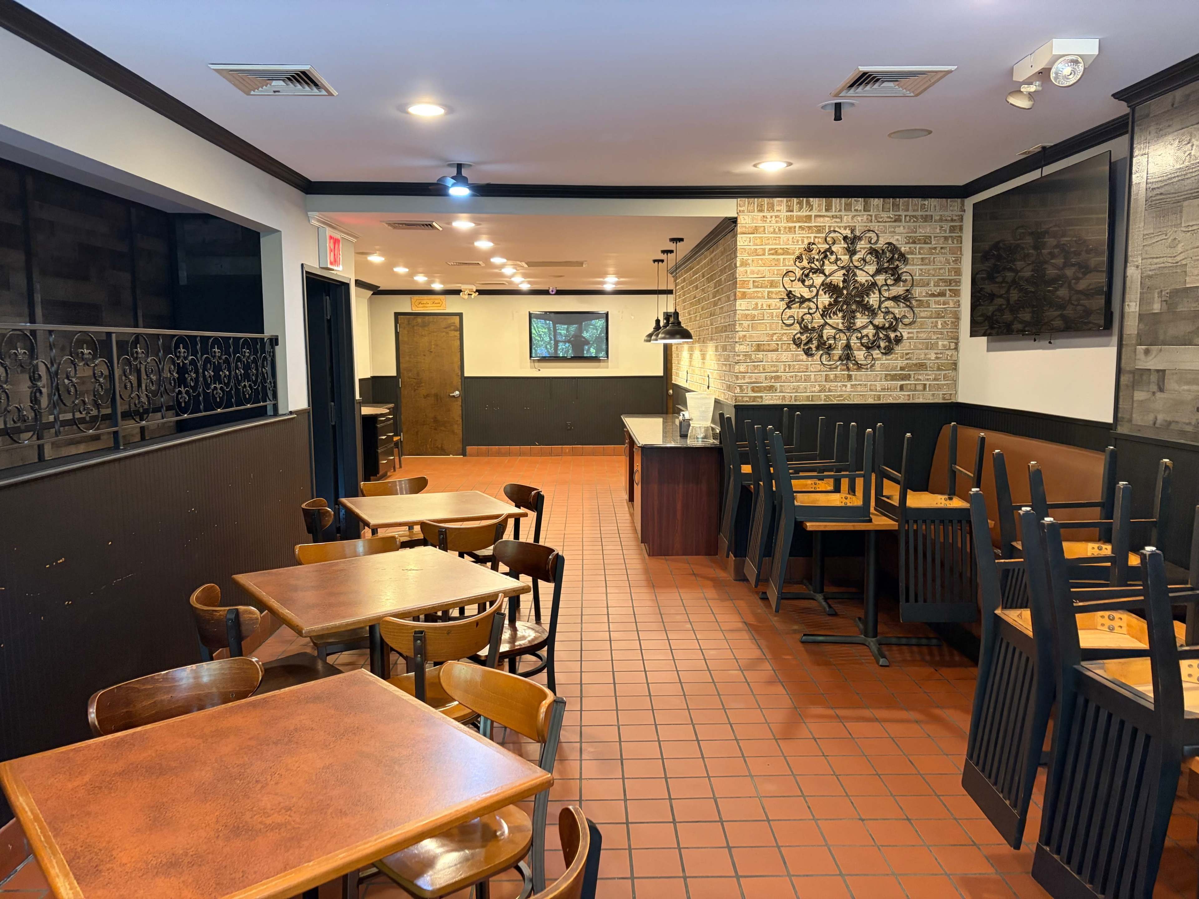 The image shows an empty restaurant interior with wooden tables and chairs stacked upside down, brick walls, and a service counter in the background.