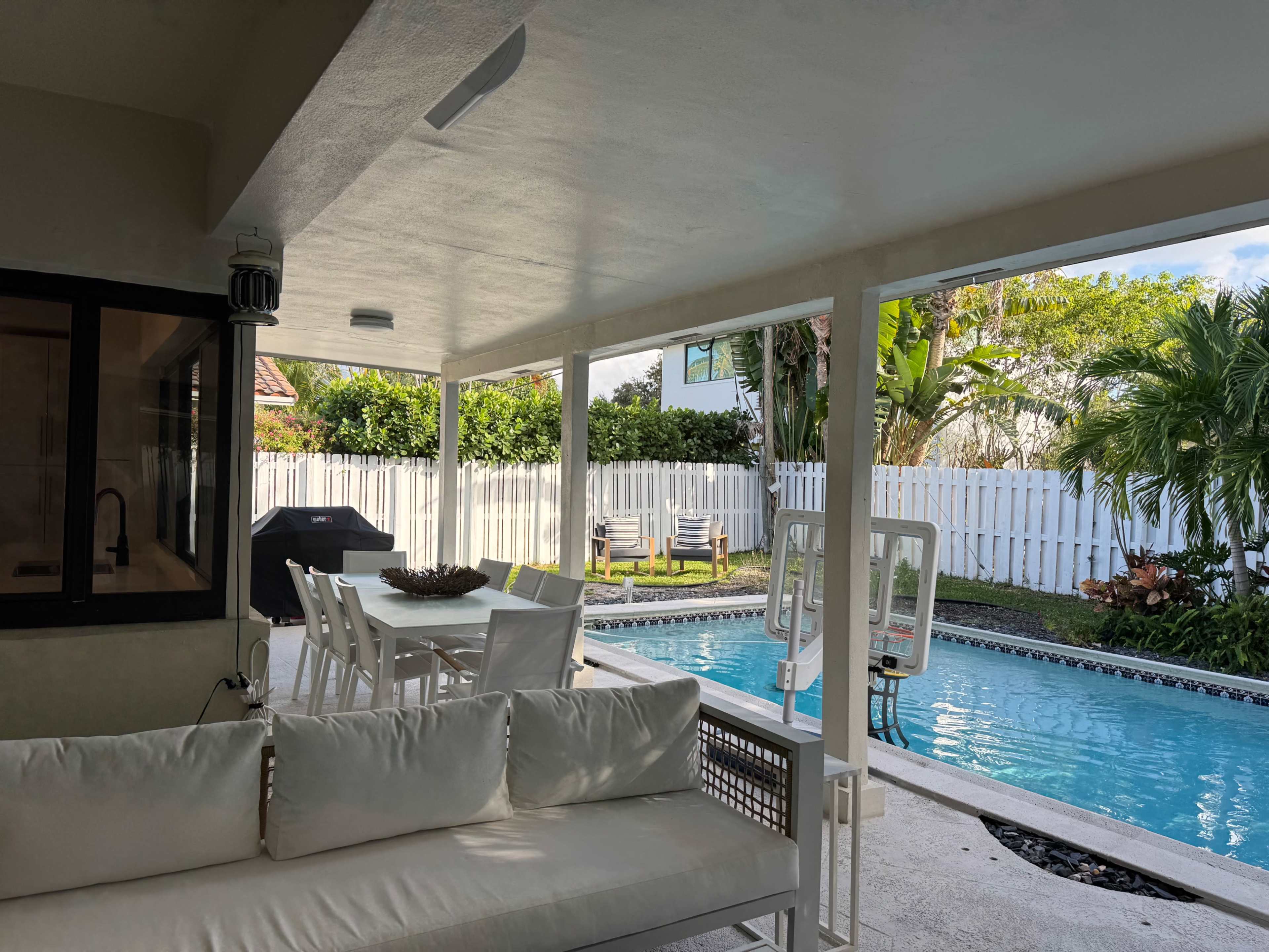 The image shows a modern outdoor patio area with a dining table and chairs beside a swimming pool, surrounded by greenery and a white fence.