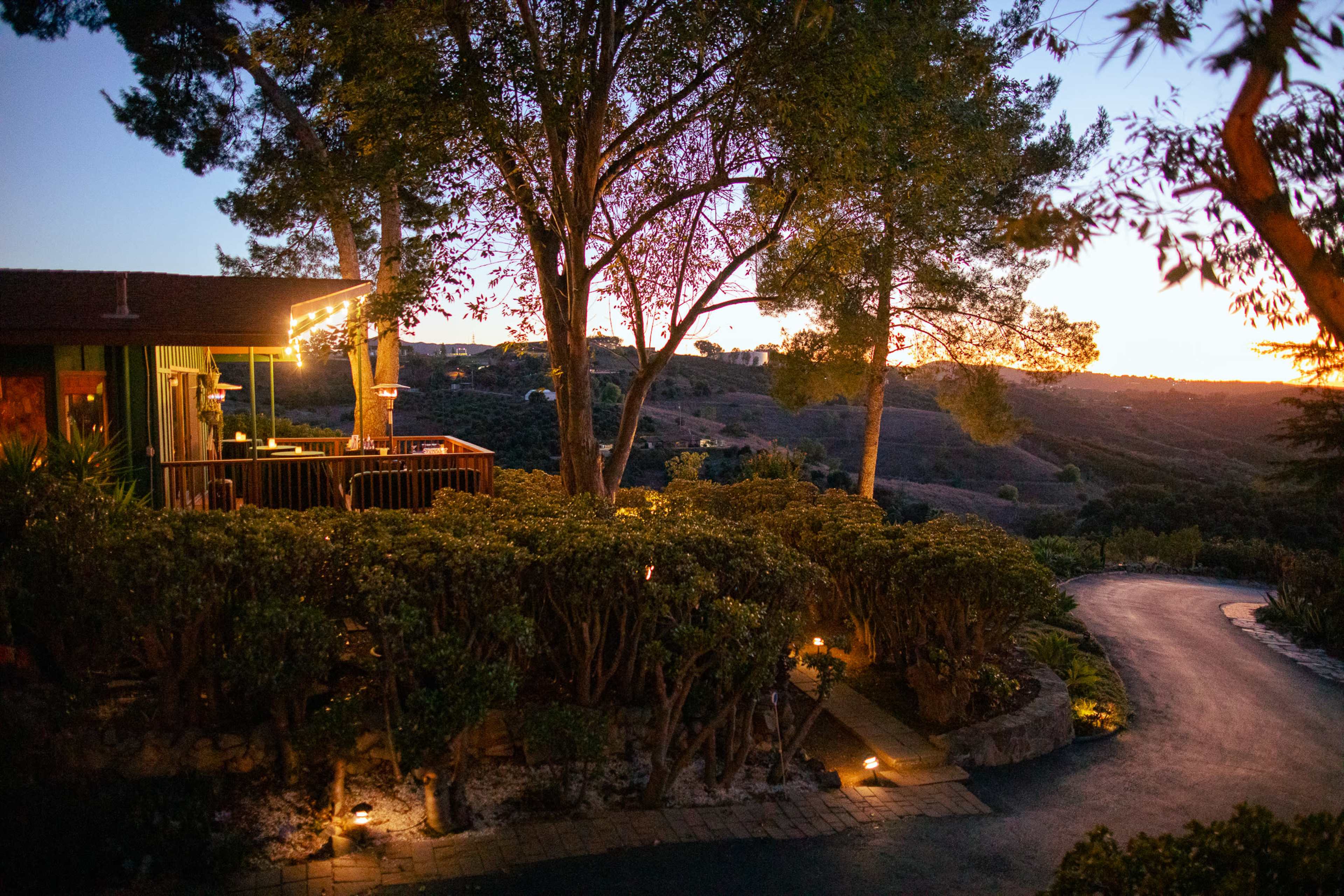 A pathway curves past a deck surrounded by trees and shrubs, leading towards rolling hills at dusk.