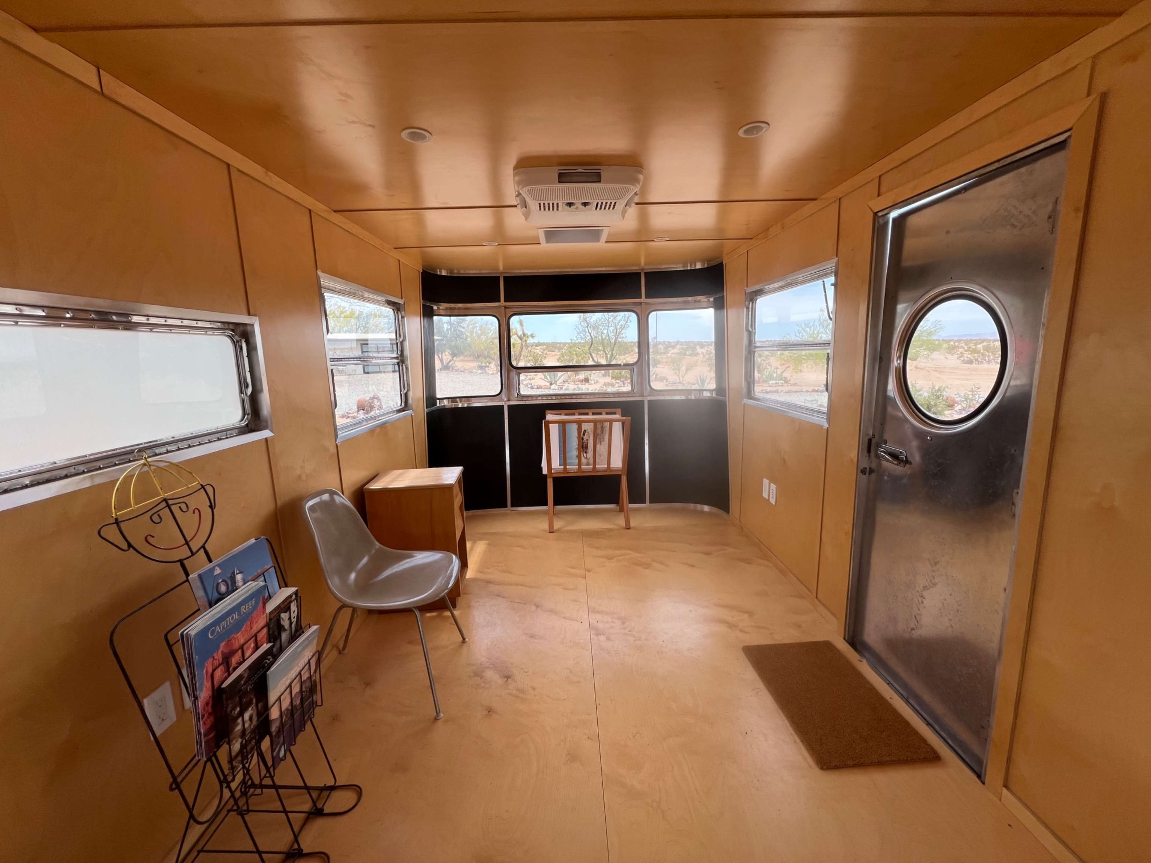 The interior of a small, minimalist trailer features wooden paneling, a chair, a table, a magazine rack, and a round windowed door.