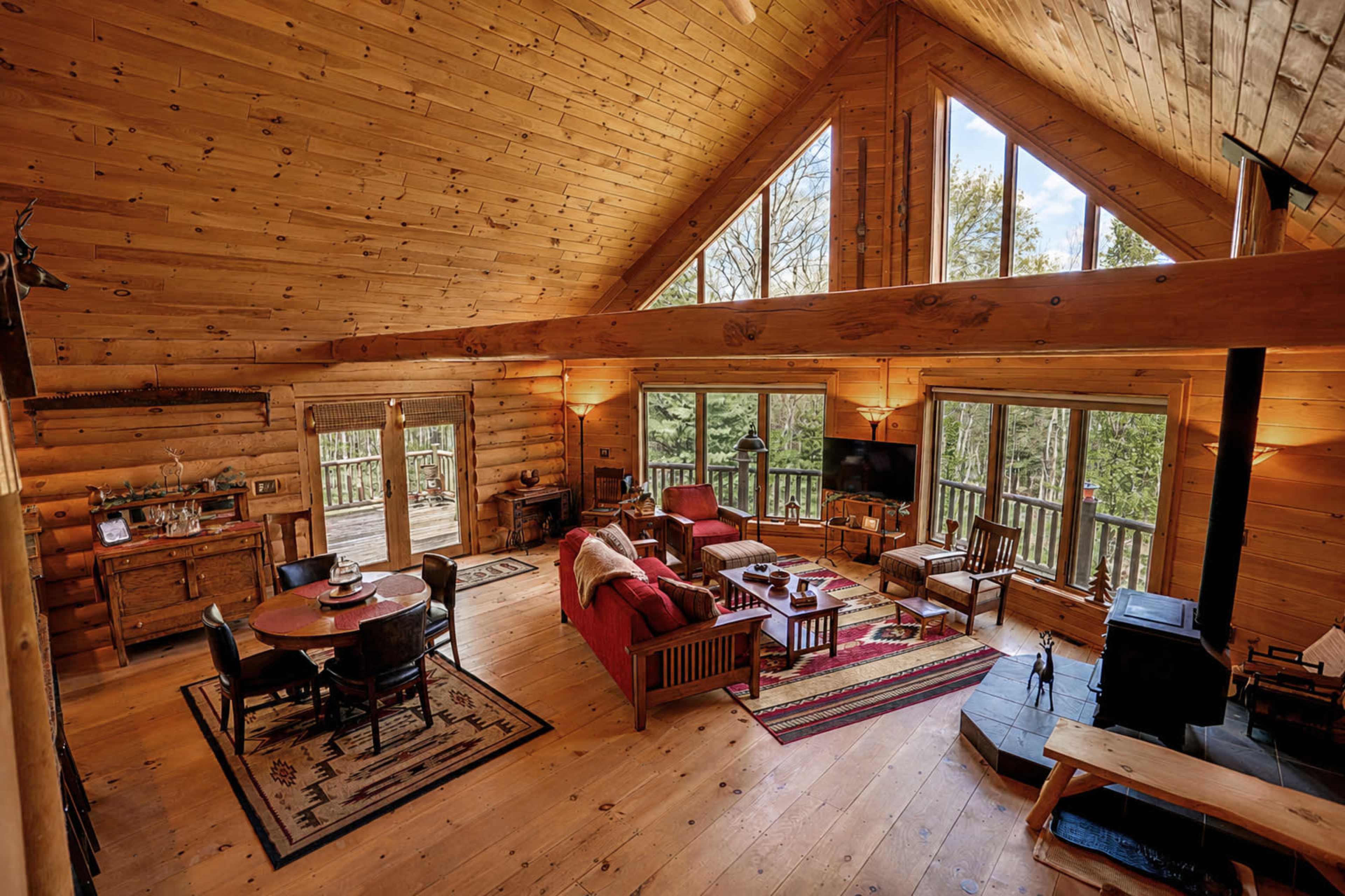 The image shows a spacious wooden cabin interior with large windows, a living area featuring red couches, a dining table, and a stone fireplace.