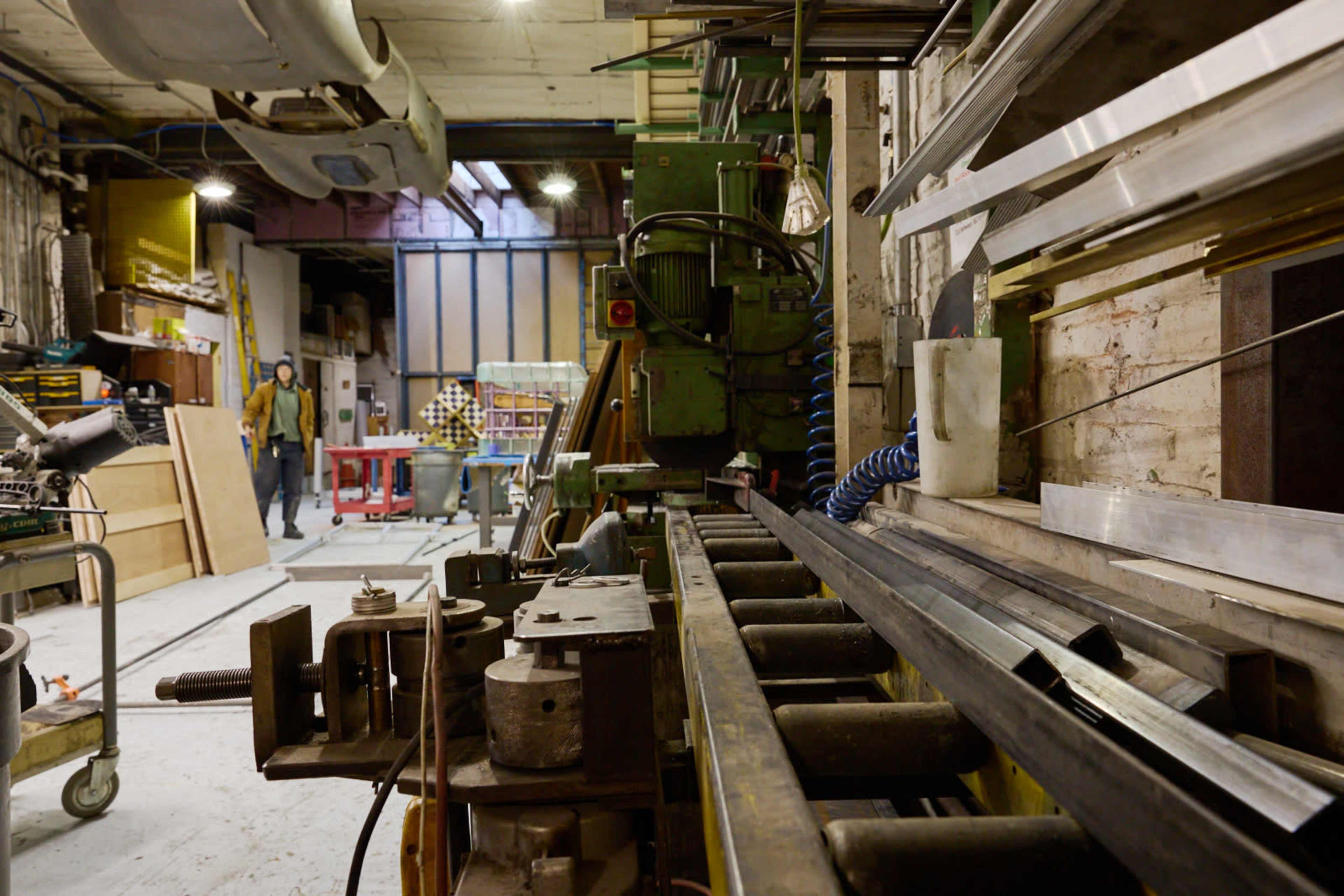 The image shows a workshop filled with various tools and machinery, featuring a metalworking machine in the foreground and another worker in the background.