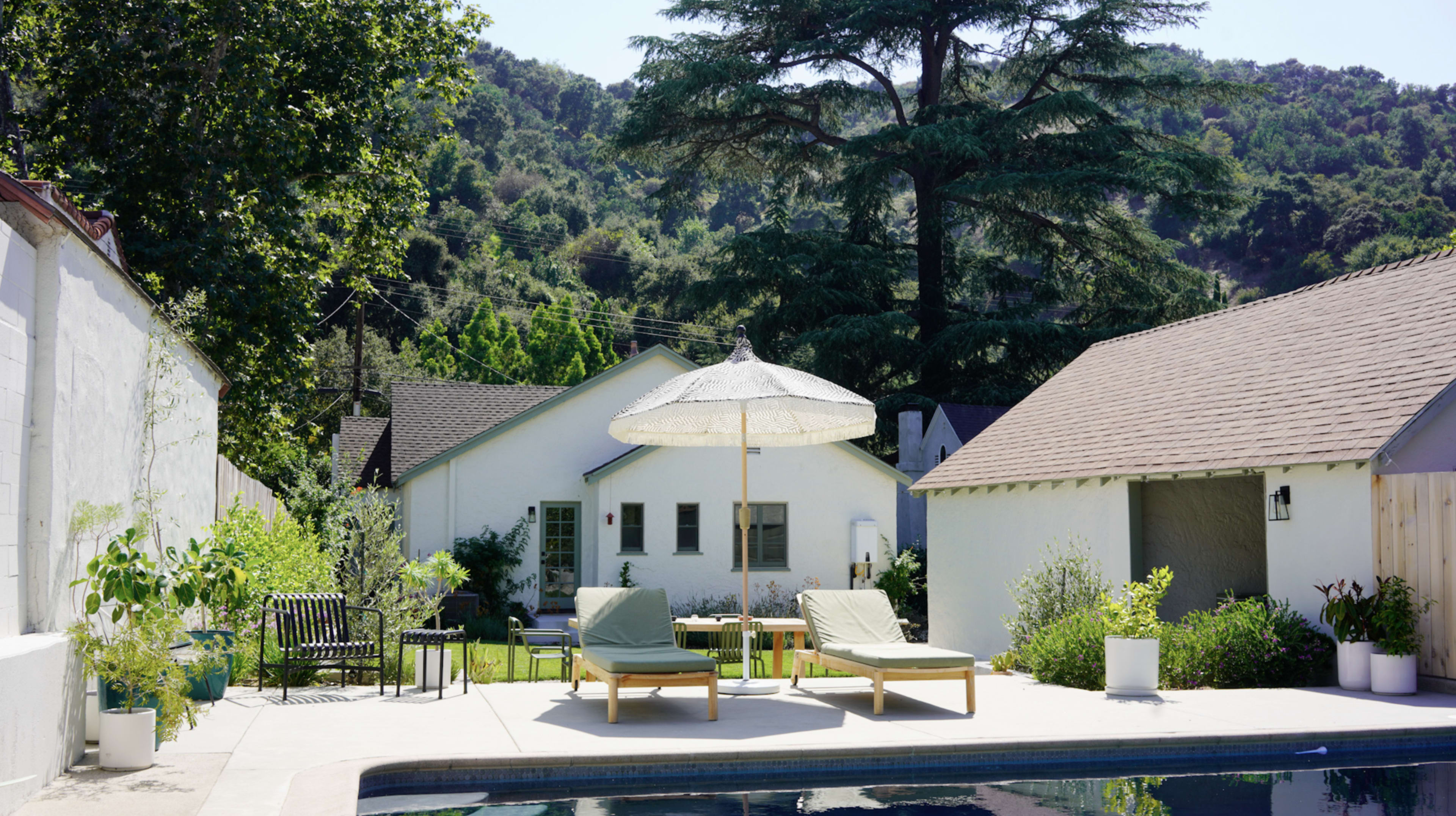 A swimming pool with two lounge chairs under a parasol is surrounded by a landscaped yard and houses against a backdrop of green hills.