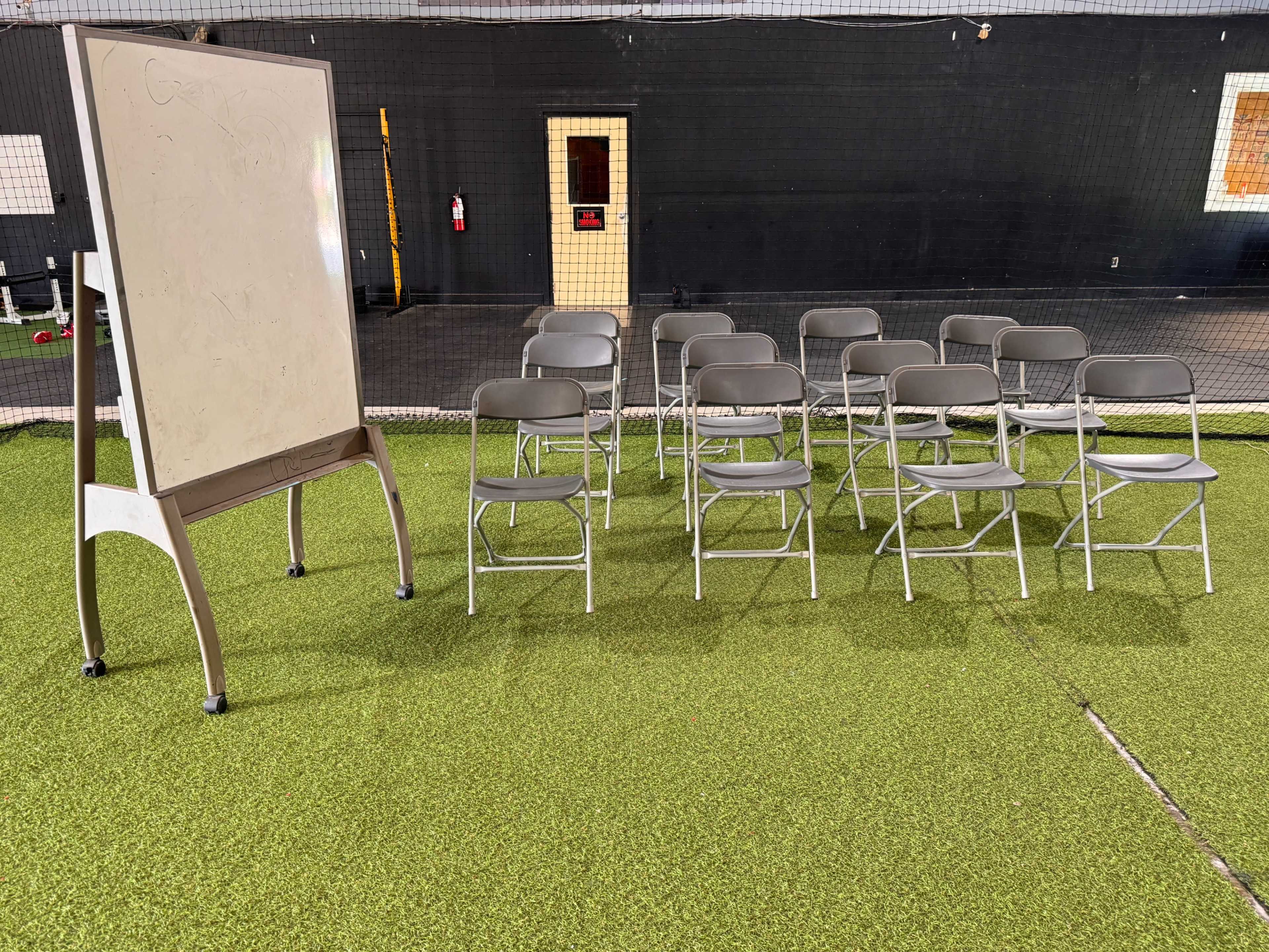 A row of gray folding chairs is arranged in front of a whiteboard in a training facility with artificial grass flooring.