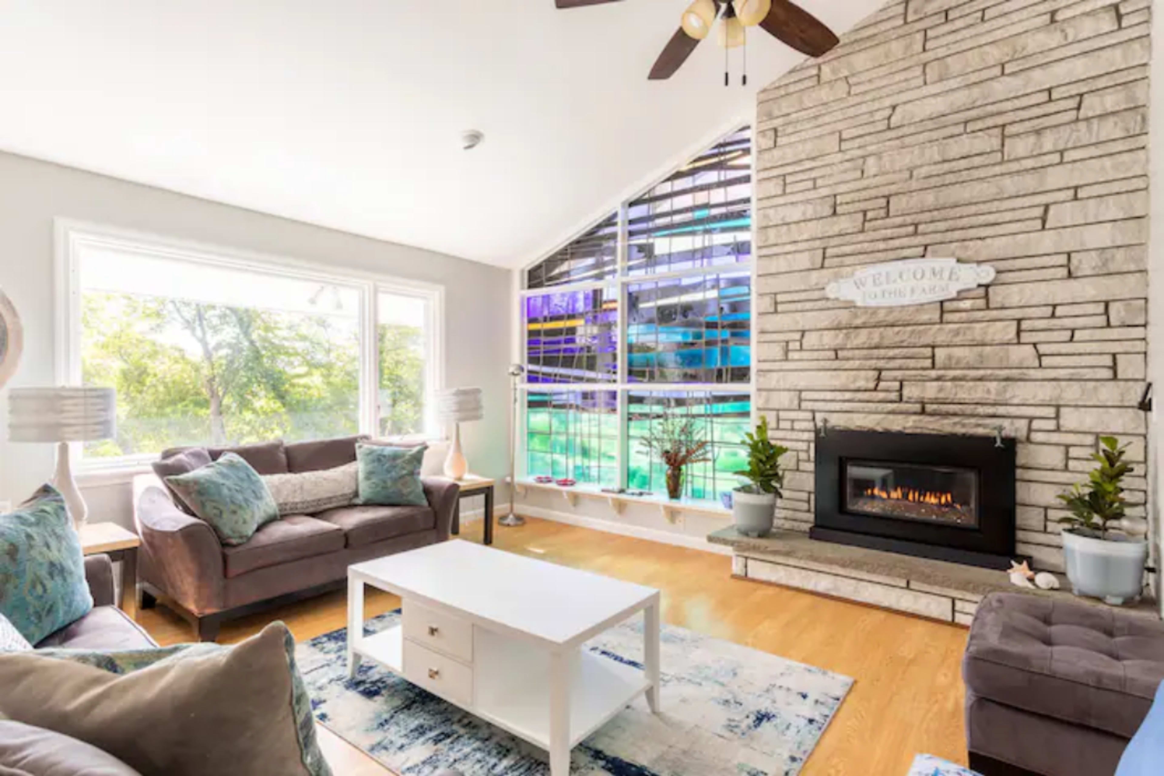 A well-lit living room features a stone fireplace, modern furniture, and large windows showcasing an outdoor view.