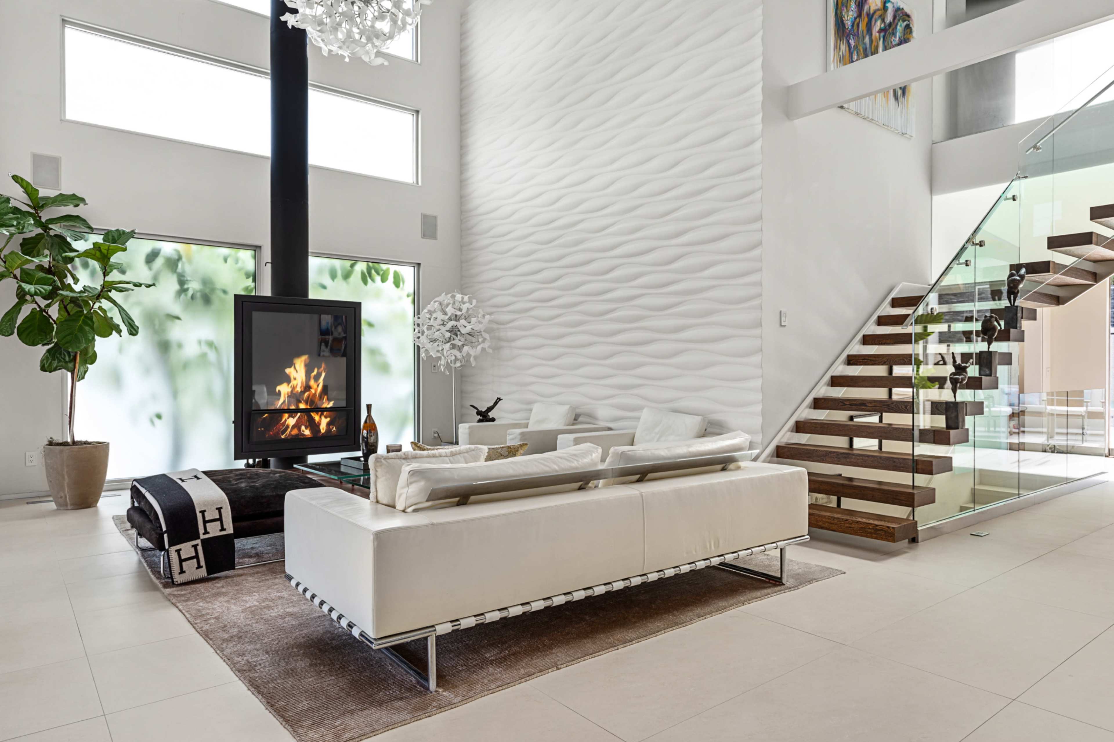 The image shows a modern living room with a white sectional sofa, a central fireplace, a large window, and a staircase leading to an upper level.